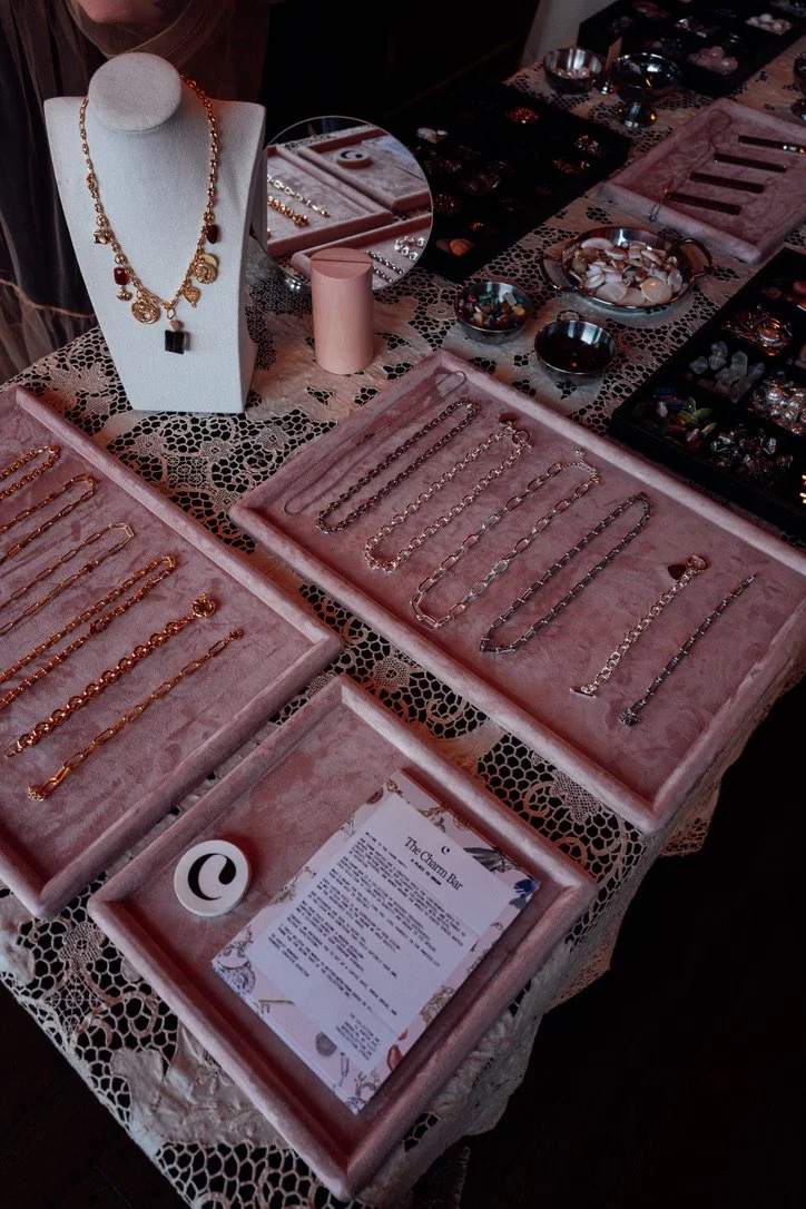 Jewelry display table with various necklaces, bracelets, and rings, featuring gold and silver pieces, a necklace on a mannequin, and a sign labeled 'The Charm Bar' in a jewelry shop.