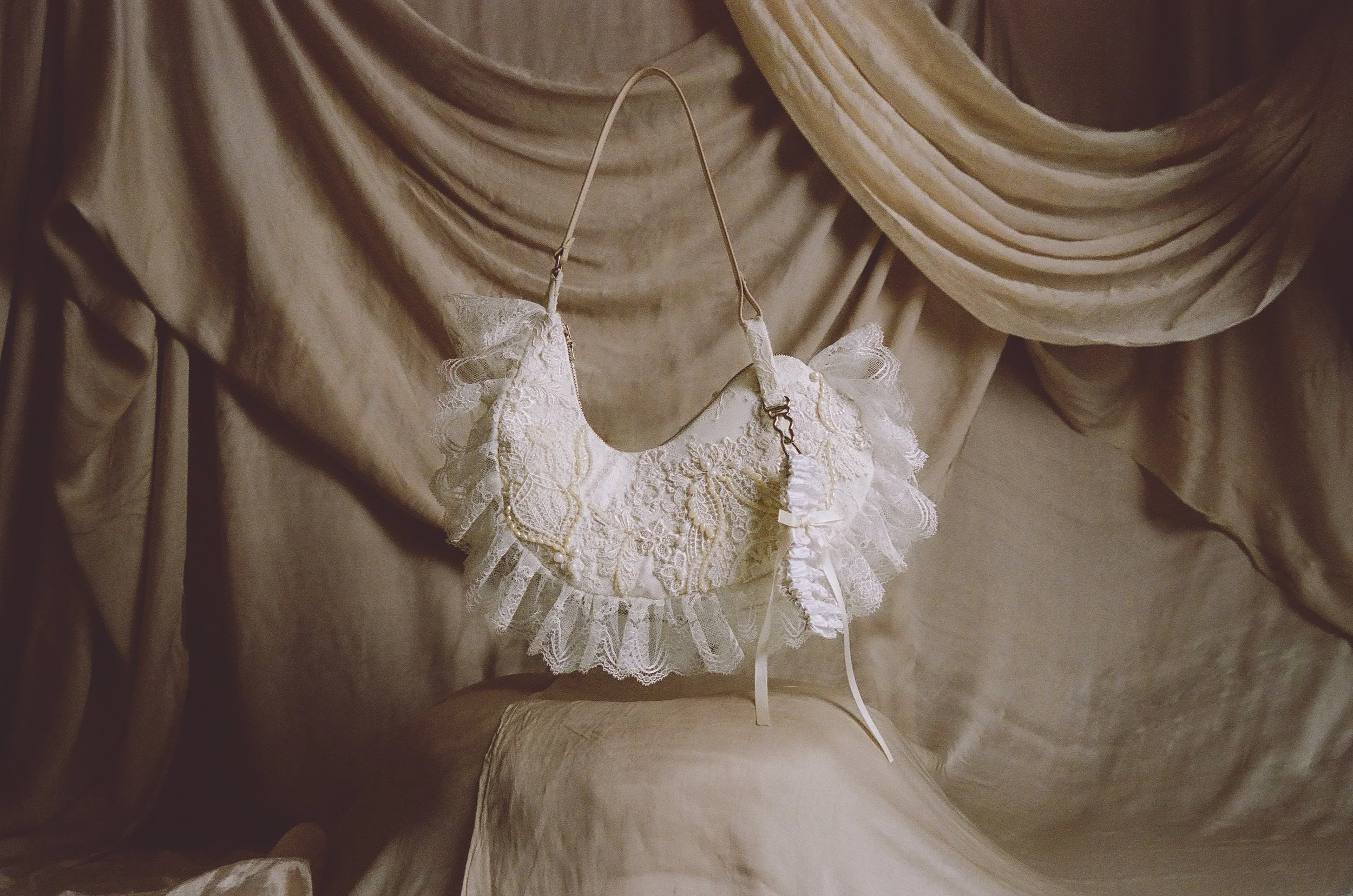 A vintage, lace-shaped handbag with ruffled lace trim is hanging against a backdrop of beige velvet drapes.