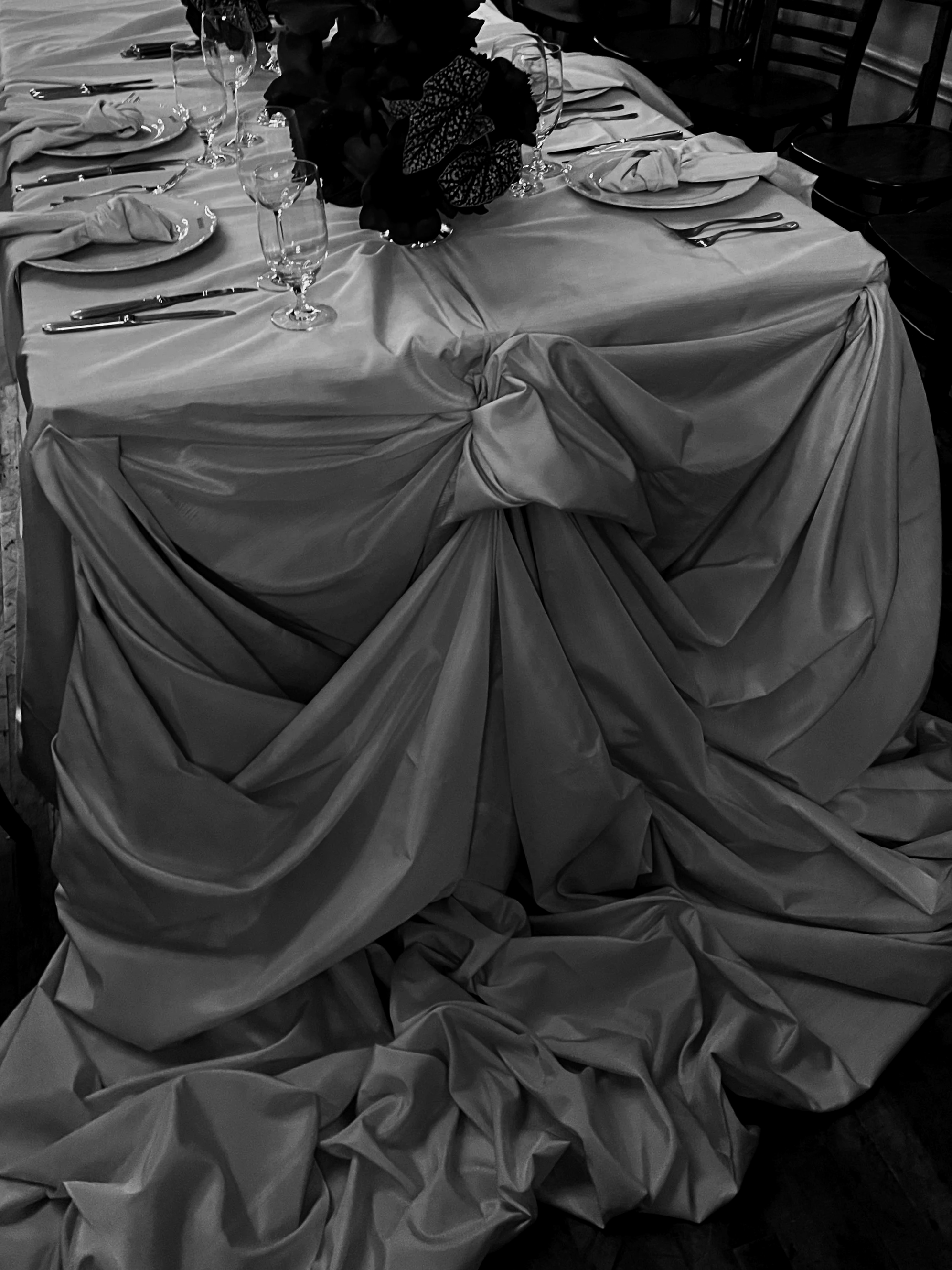 A decorated banquet table with a draped tablecloth, set with plates, napkins, forks, knives, wine glasses, and a floral centerpiece in a black-and-white photo.