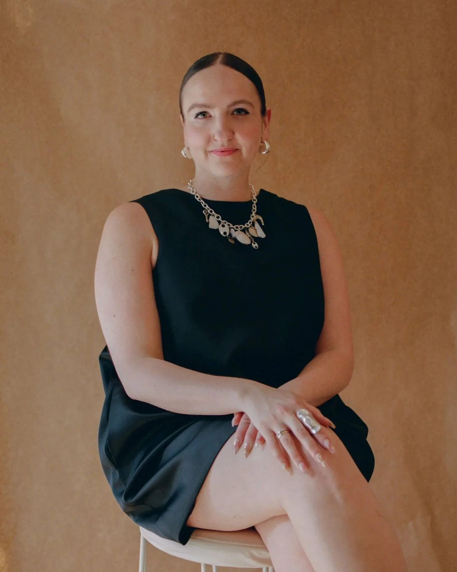 A woman with dark hair tied back, wearing a black sleeveless top and black skirt, sitting on a beige chair against a brown background. She has earrings, a chunky silver necklace, and rings on her fingers. She is smiling slightly and has her legs crossed, with her hands resting on her knee.