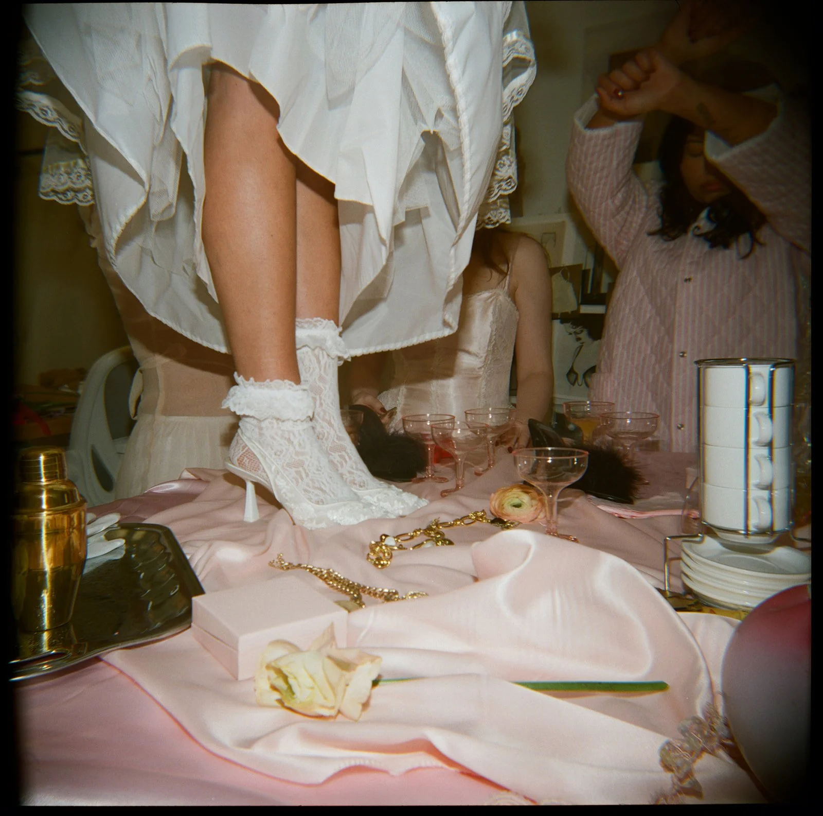 Close-up of a table with pink satin cloth, jewelry, glasses, and a flower. A person in white lace gloves and vintage dress is standing on the table. Other women are in the background, dressed vintage-style, with one covering her face with her arm.