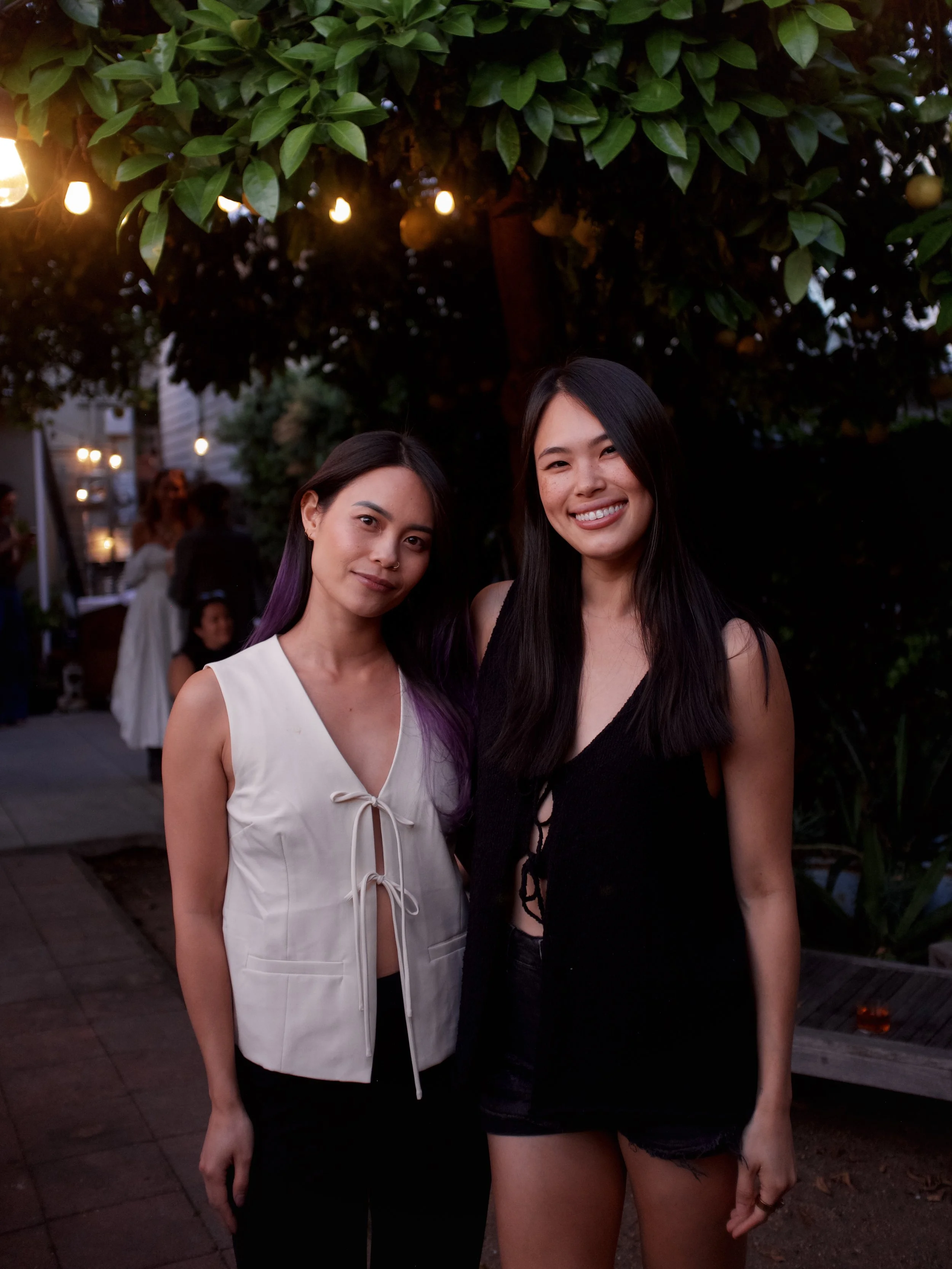Two women standing outdoors in front of a tree with hanging lights at dusk, smiling at the camera.