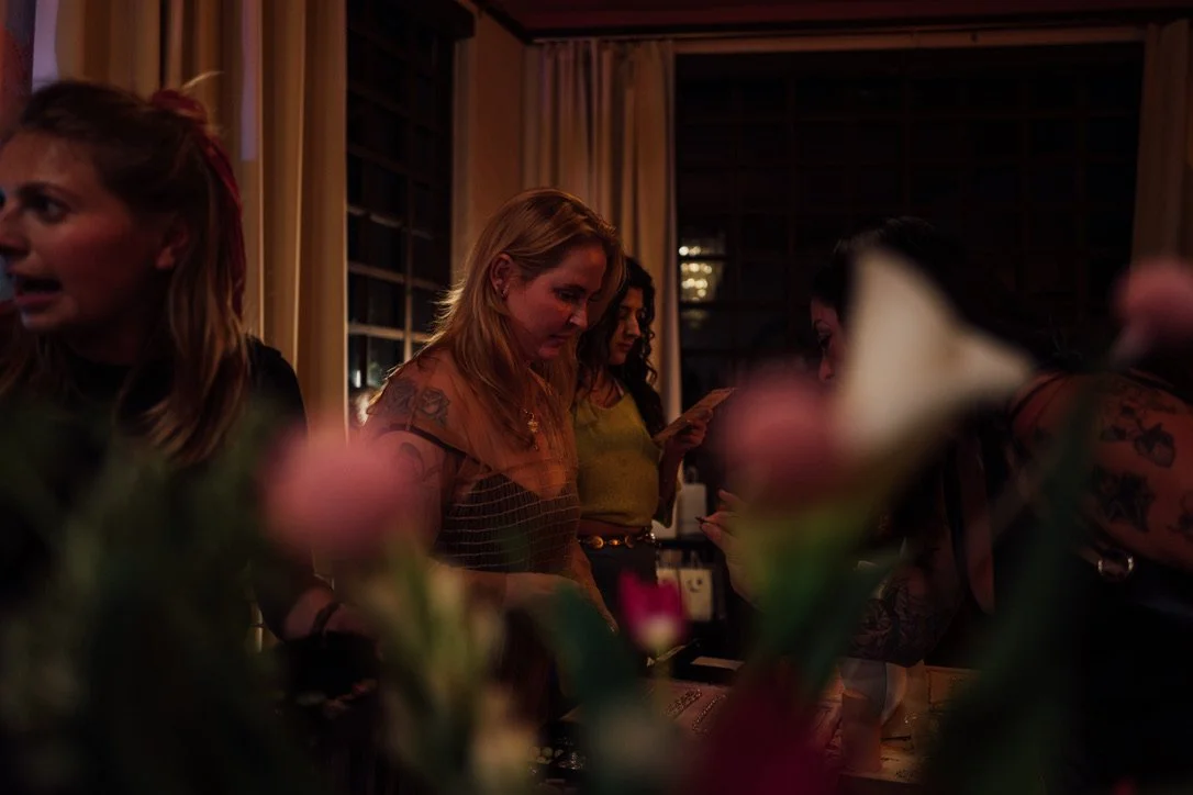 Women gathered around a table at a social event, with flowers in the foreground and warm indoor lighting.