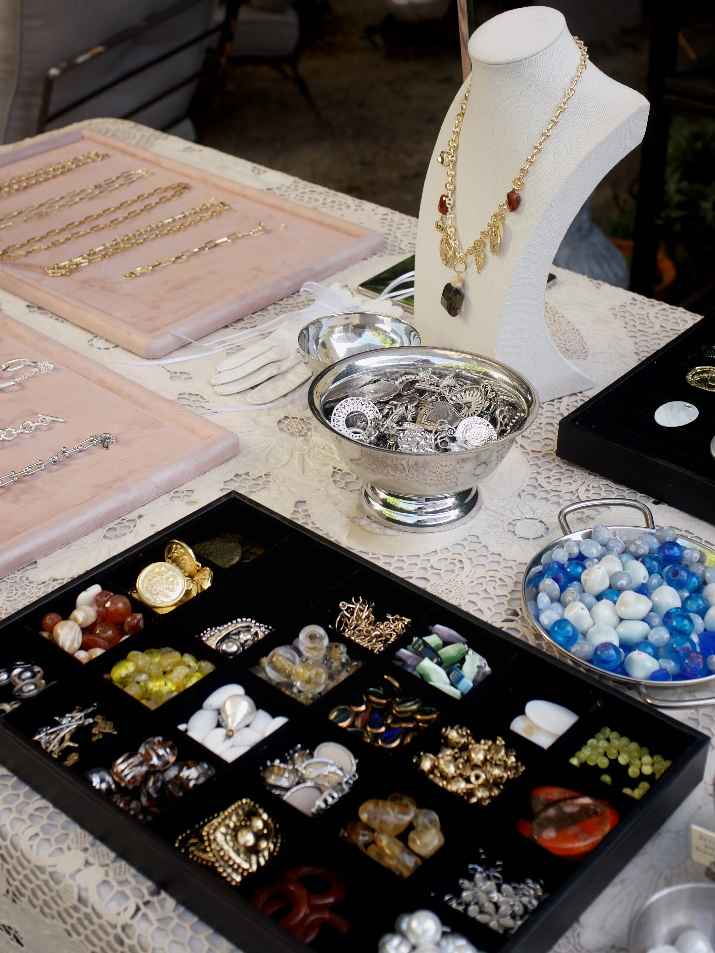 Various jewelry items including necklaces, bracelets, rings, and earrings displayed on trays and a mannequin bust, some made of gold and others of beads, with a black jewelry box on the side and a silver bowl filled with jewelry parts.