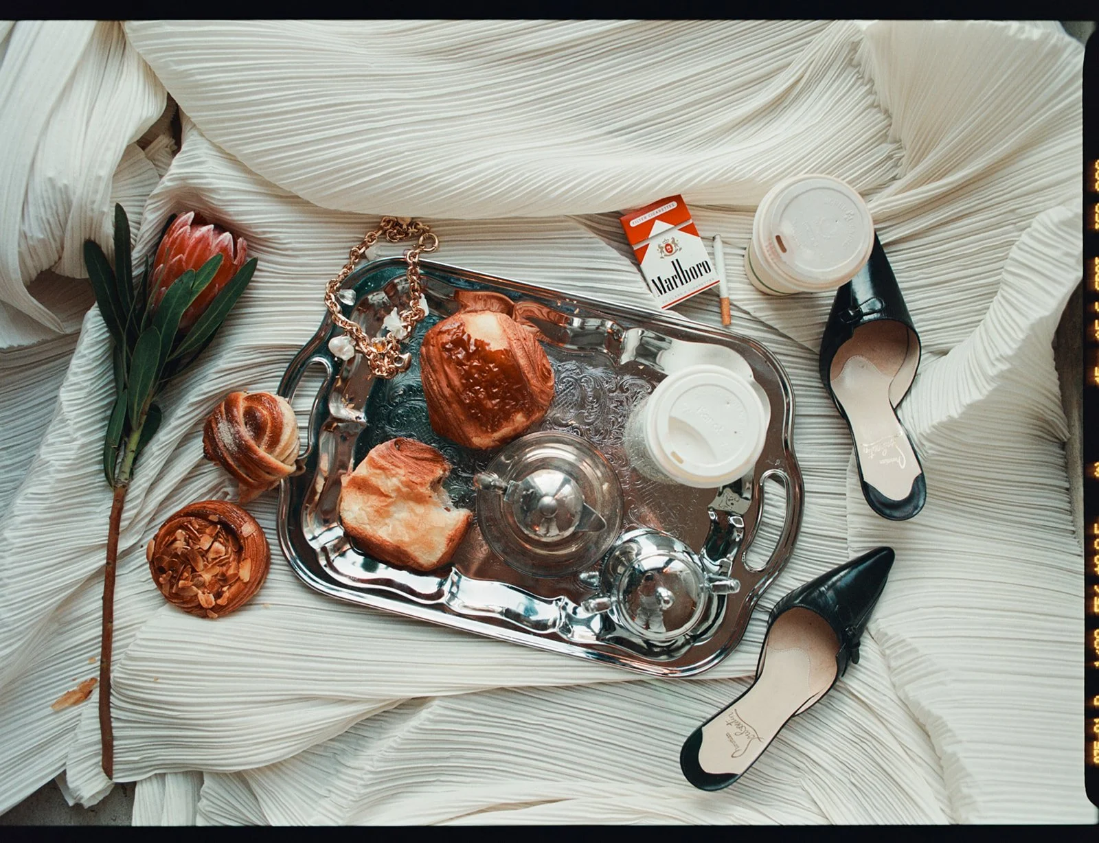 A silver tray with pastries, a pearl necklace, two disposable coffee cups, a pack of Marlboro cigarettes, a lipstick, a pair of black high heels, and some green leaves arranged on a white fabric surface.
