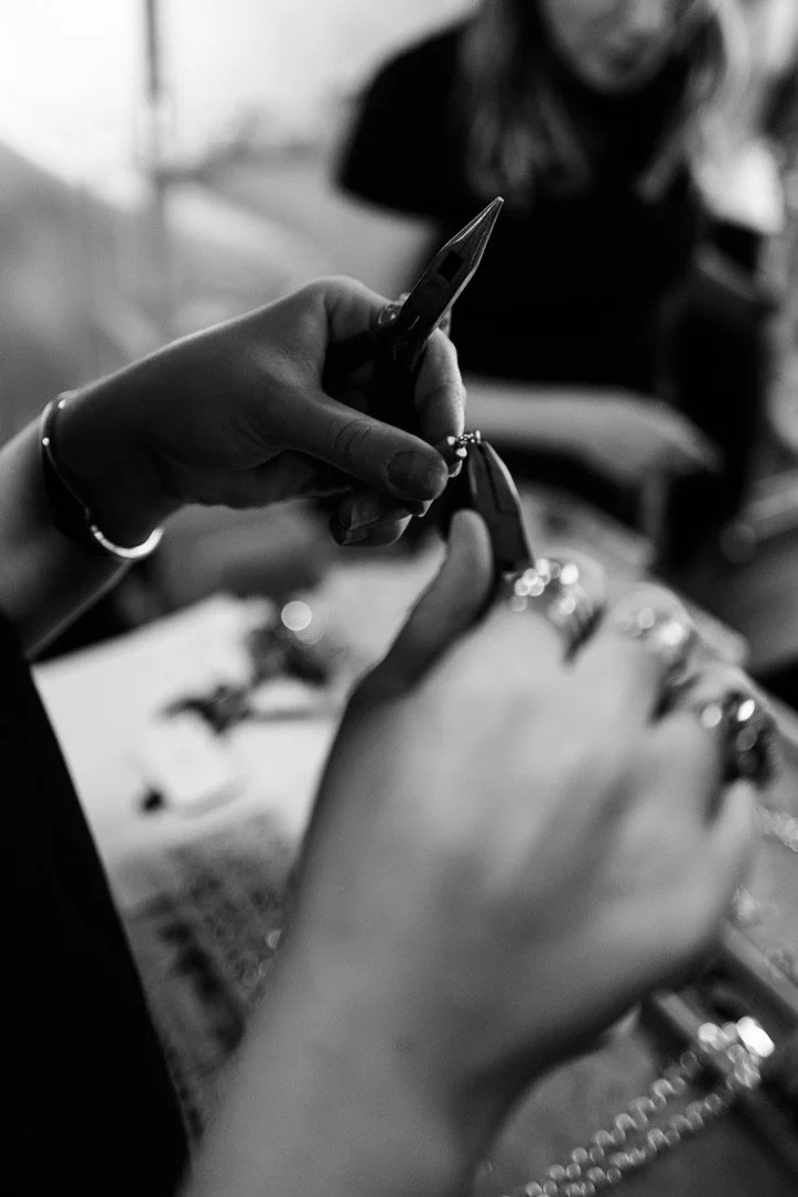 Close-up of hands with rings holding jewelry pliers, working on jewelry, with blurred person in background.