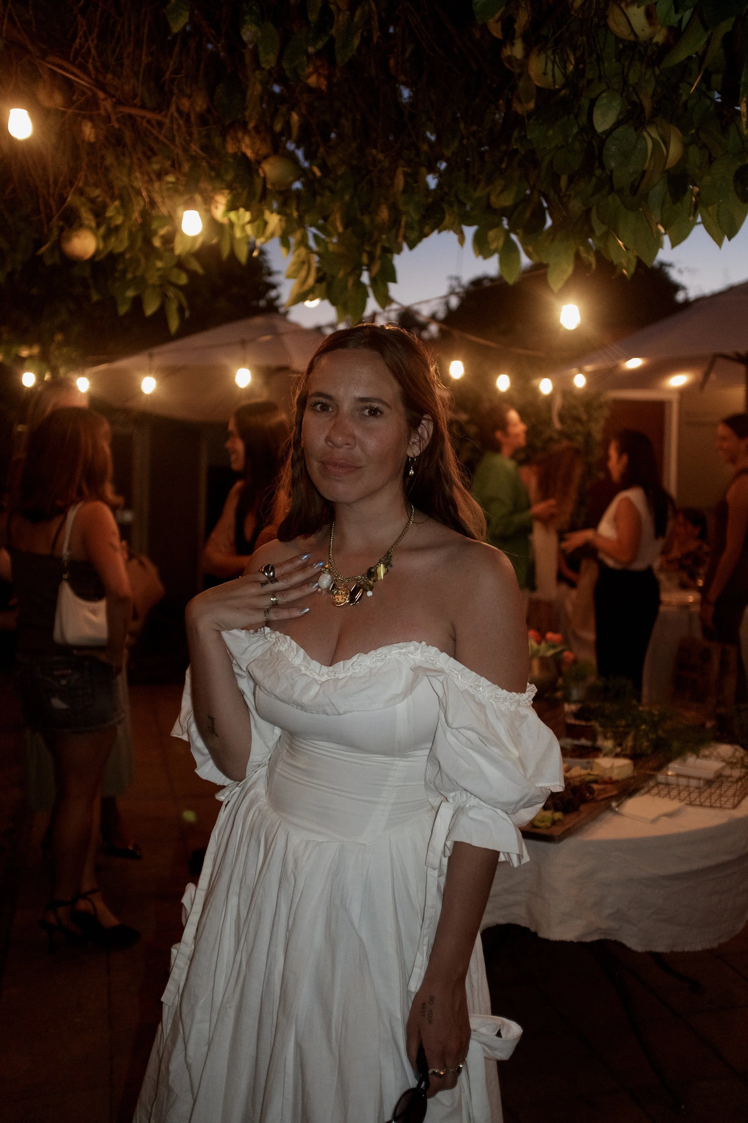 A woman in a white off-shoulder dress smiling at an outdoor evening gathering, with string lights and other people in the background.