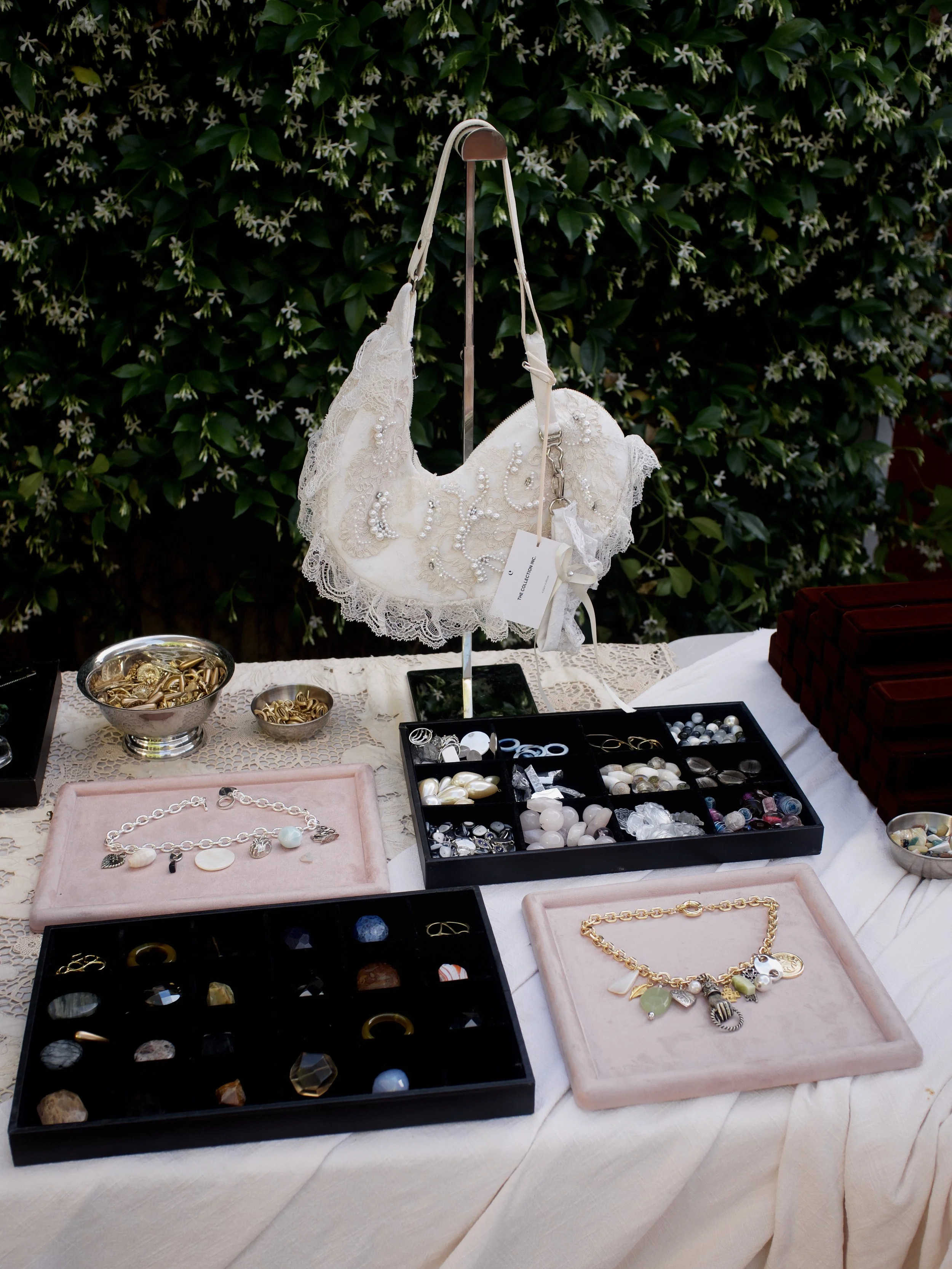 A display of jewelry including necklaces, bracelets, and rings arranged on a table with a white tablecloth, some in velvet trays and others in bowls, set against a background of green leafy plants.