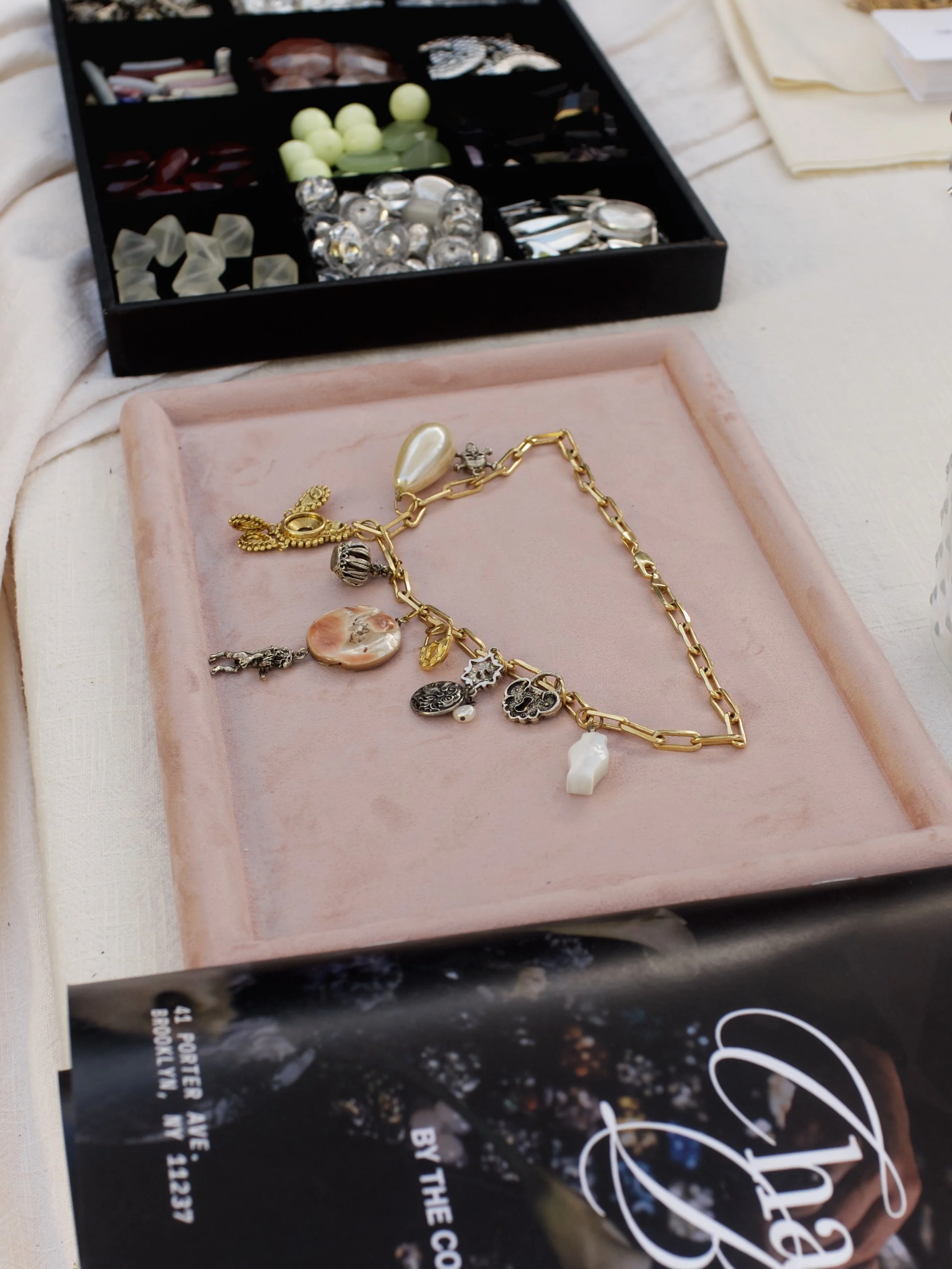Close-up of jewelry, including a gold chain necklace with various charms and large pendant, placed on a pink velvet display tray. In the background, a black jewelry box with compartments containing various loose beads, gemstones, and jewelry componen