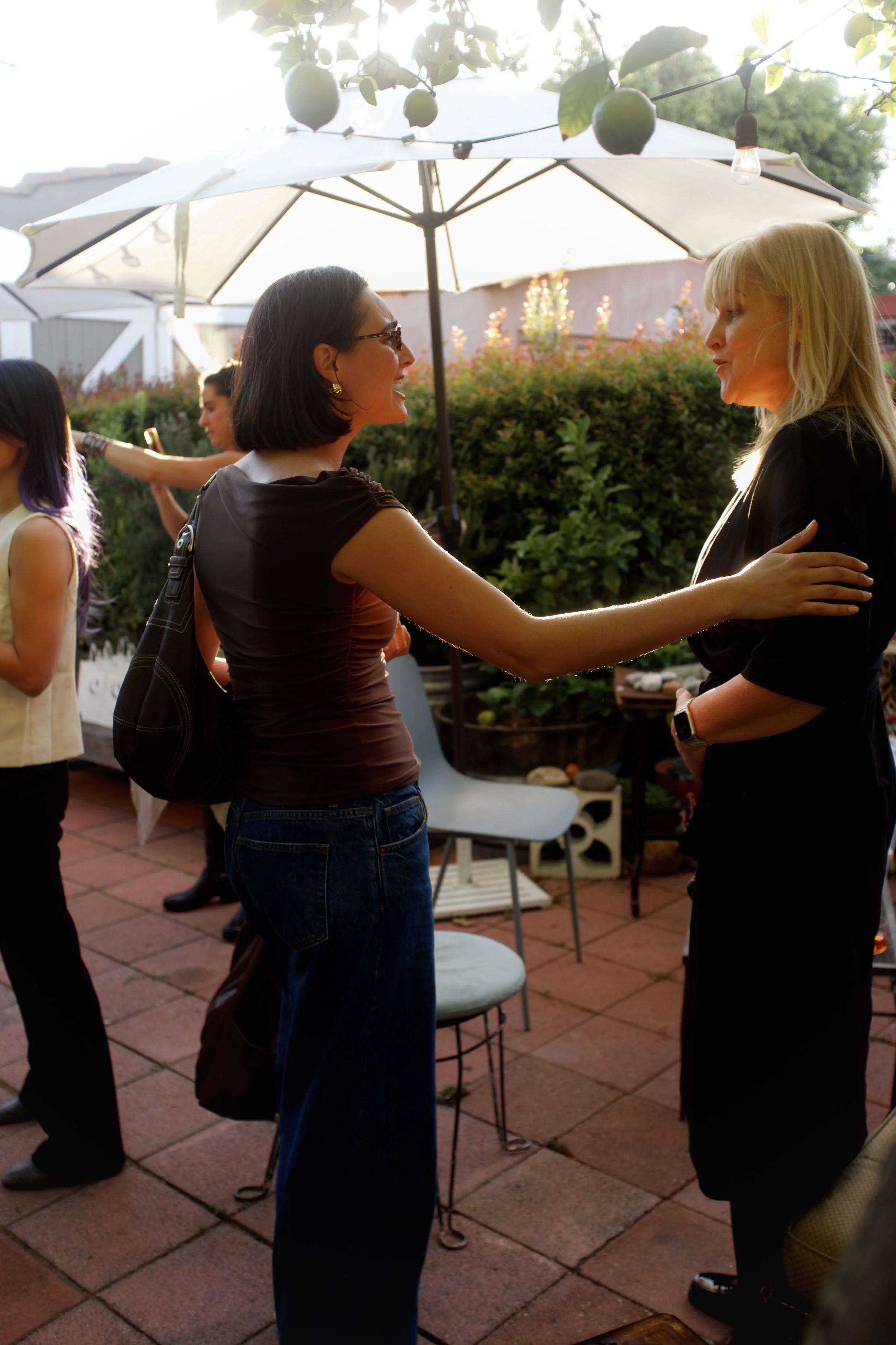 Two women are greeting each other at an outdoor gathering, with a lush garden and patio furniture, under large white umbrellas, during the evening with string lights.