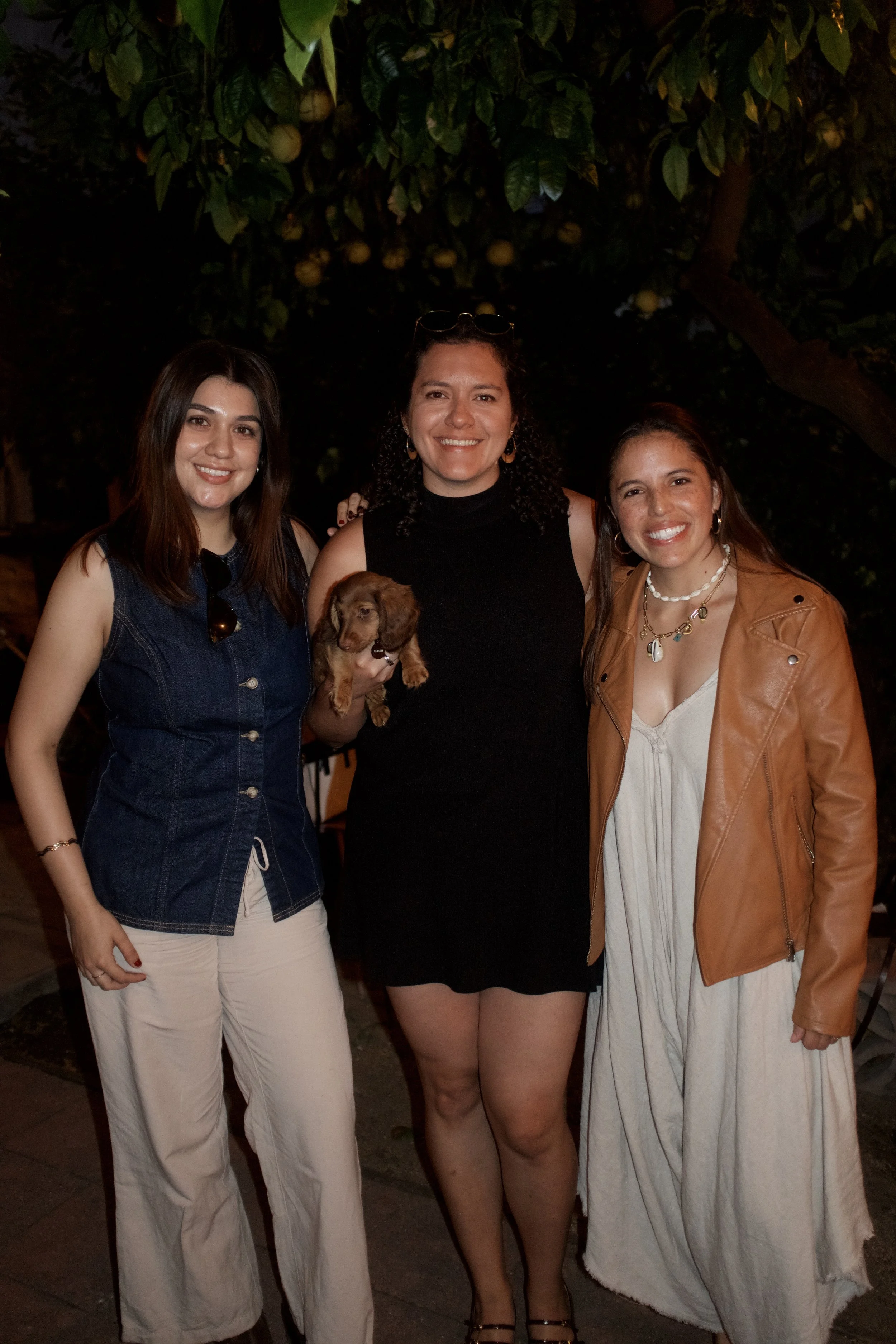 Three women standing together outdoors at night, smiling, with a tree bearing fruit in the background, and a small dog being held by the woman in the middle.