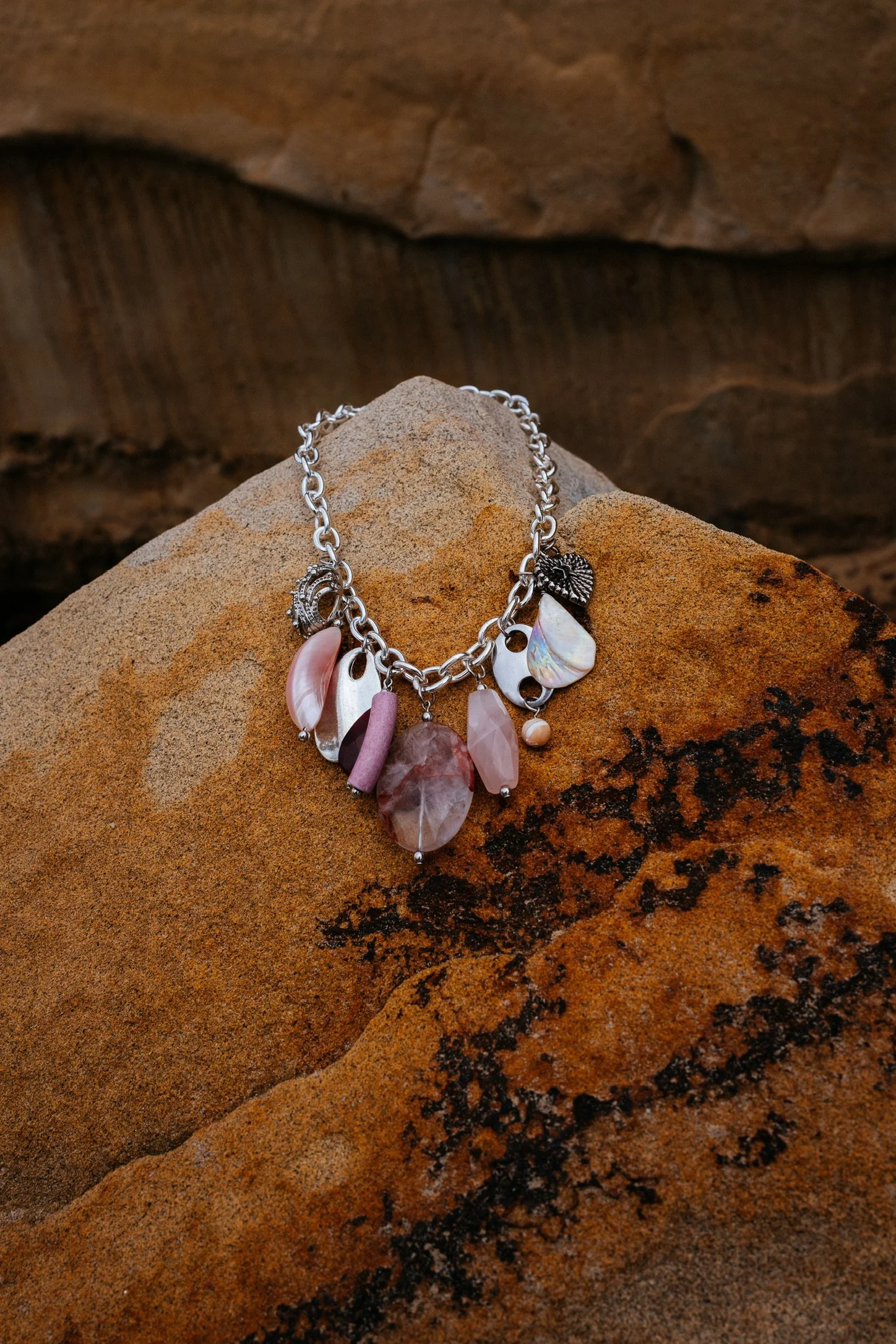 A silver charm bracelet with various shell and stone charms resting on an orange-brown rock surface.