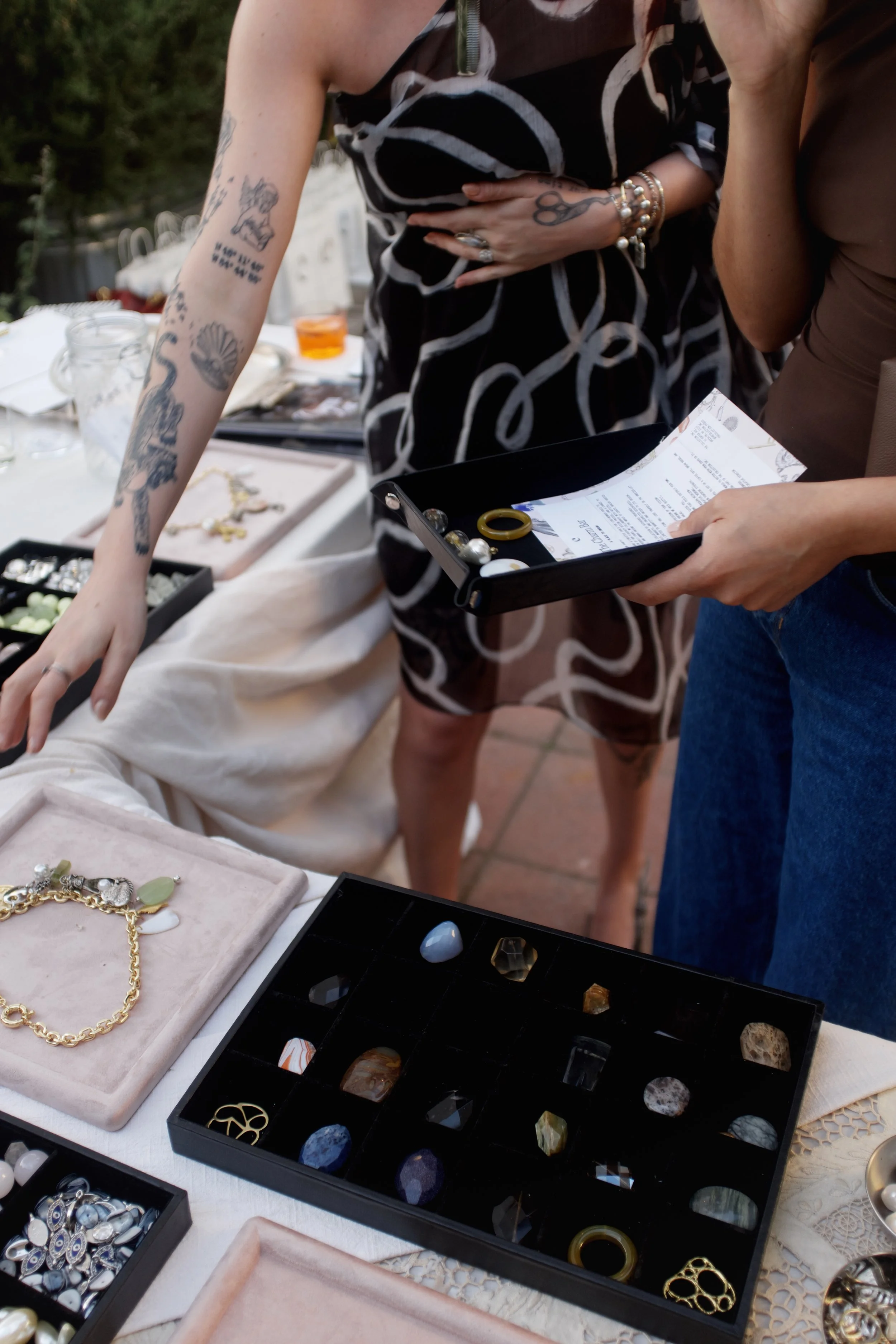 People browsing jewelry at an outdoor market table with various necklaces, rings, and gemstones on display.