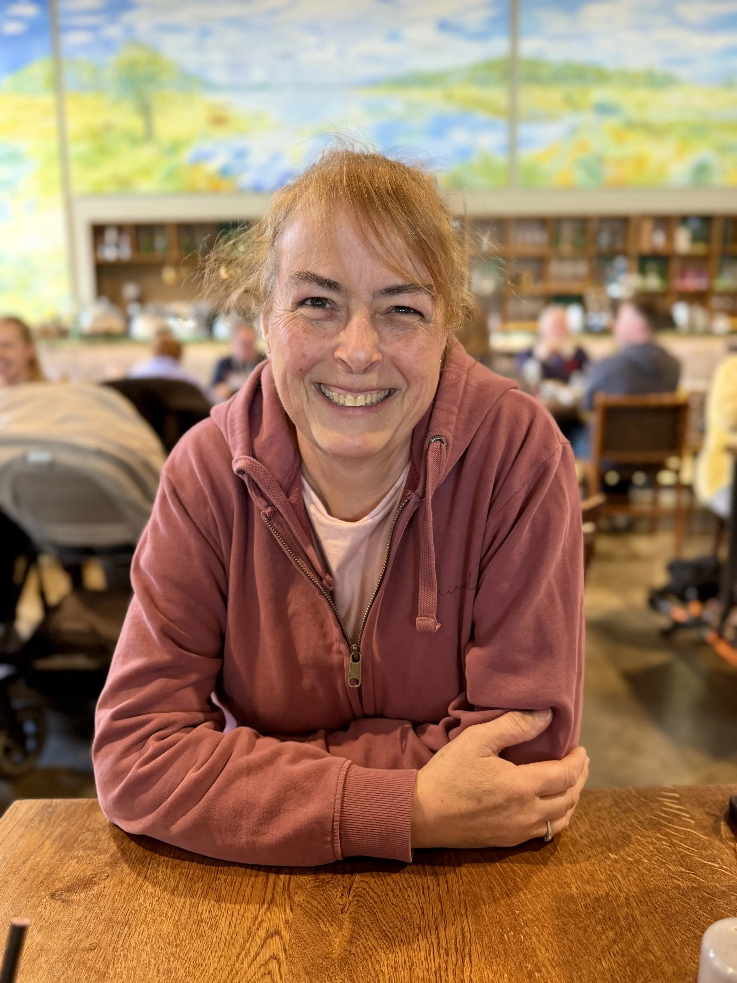 A smiling woman with reddish hair wearing a pink hoodie, sitting at a wooden table in a restaurant or cafe with a blurred background of other people and colorful artwork.