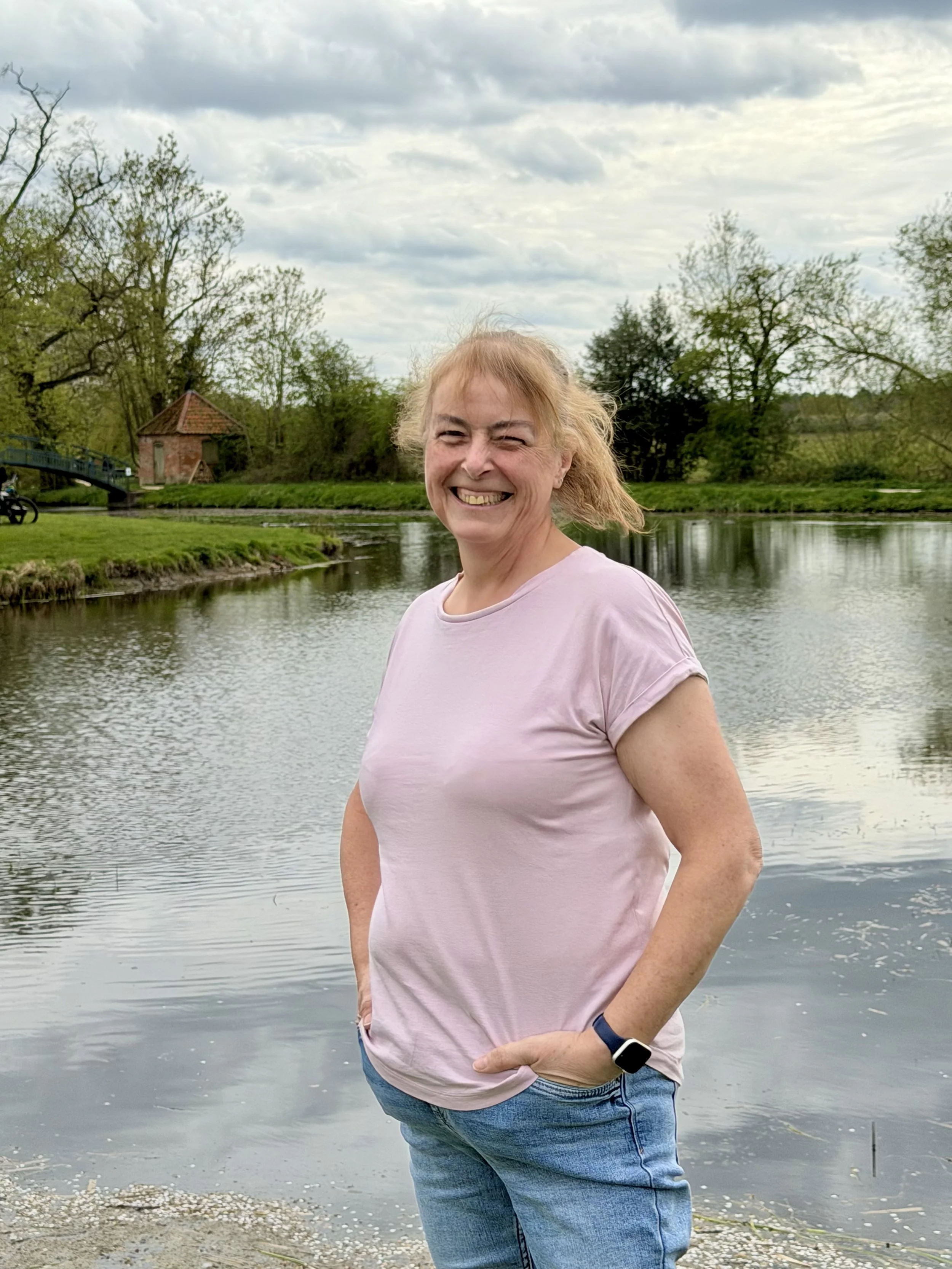 A smiling woman with short blonde hair standing near a river in a park, wearing a pink t-shirt and blue jeans, with trees and a small brick building in the background under a cloudy sky.
