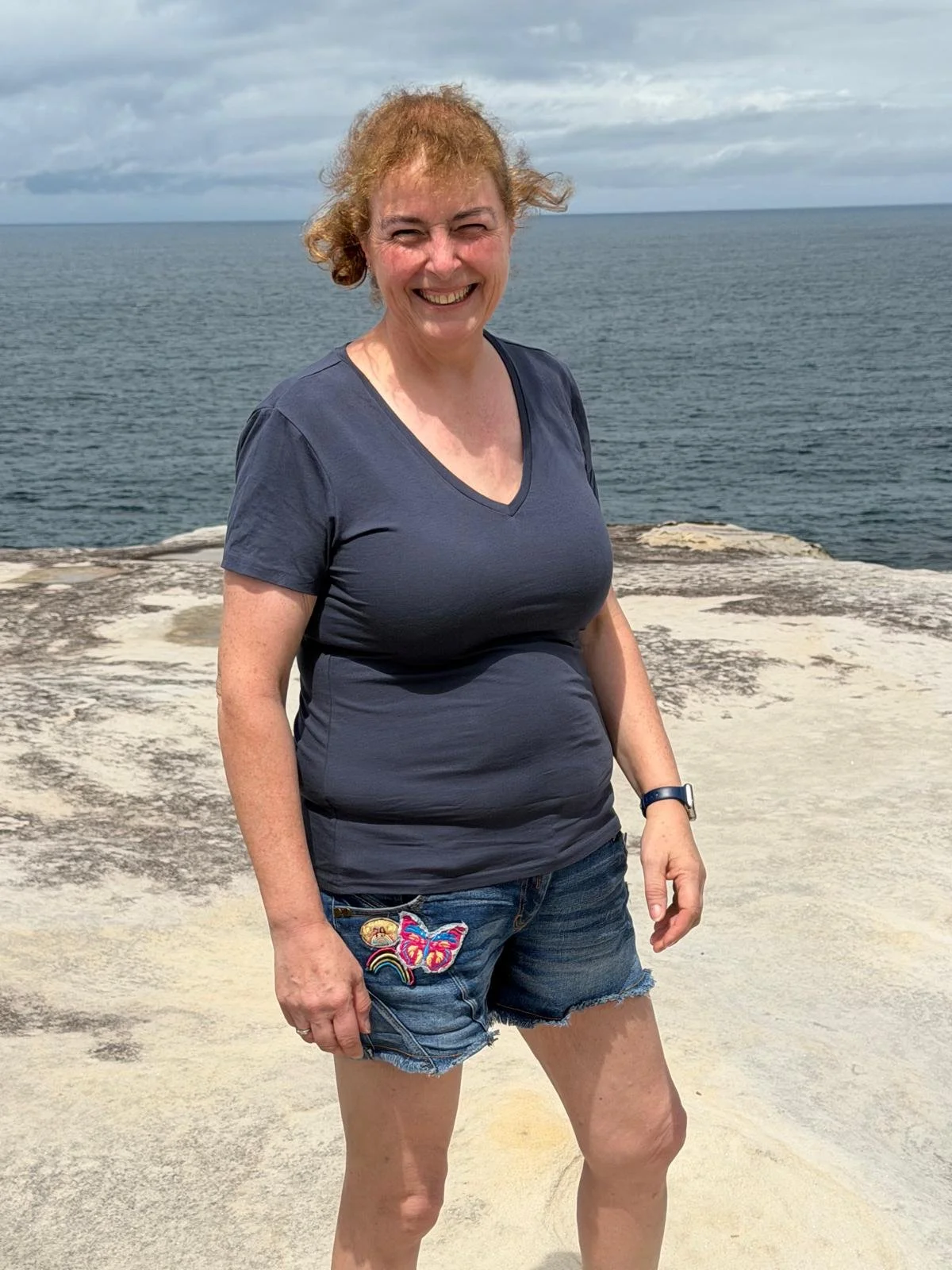 A woman smiling outdoors near the ocean with cloudy skies in the background, wearing a navy blue t-shirt and denim shorts decorated with patches.