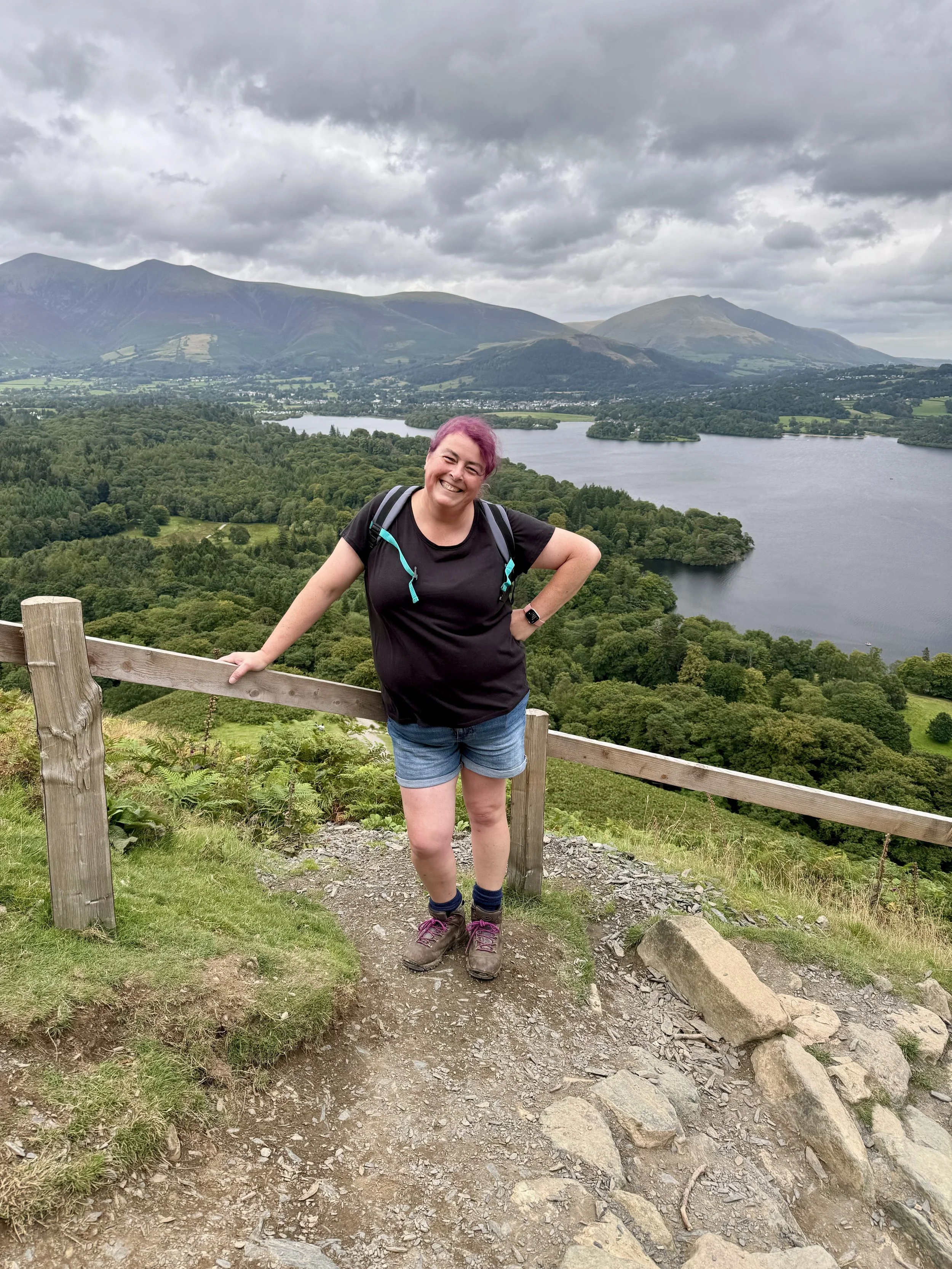 A woman with pink hair smiling and posing on a hiking trail overlooking a lake and mountains in the distance under a cloudy sky.