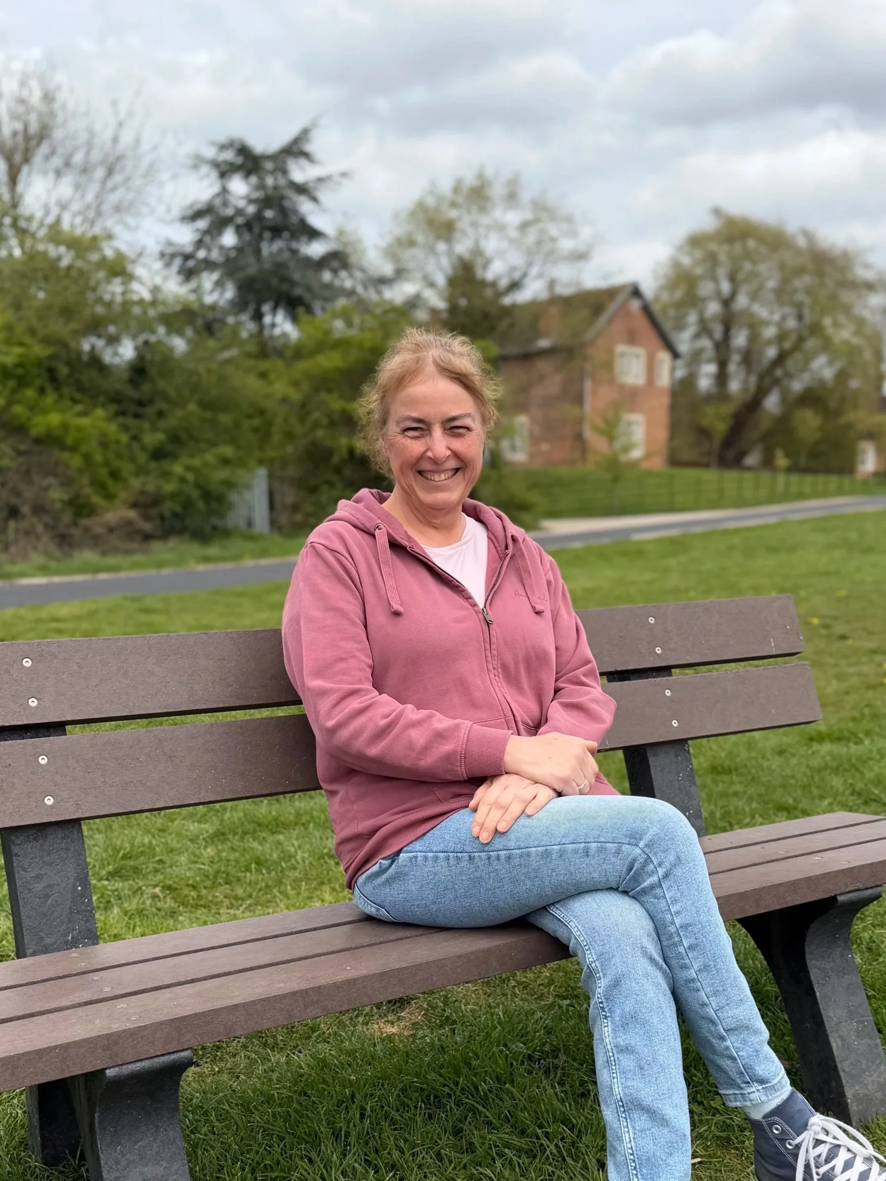 A woman smiling and sitting on a park bench outdoors with green grass, trees, and a house in the background.