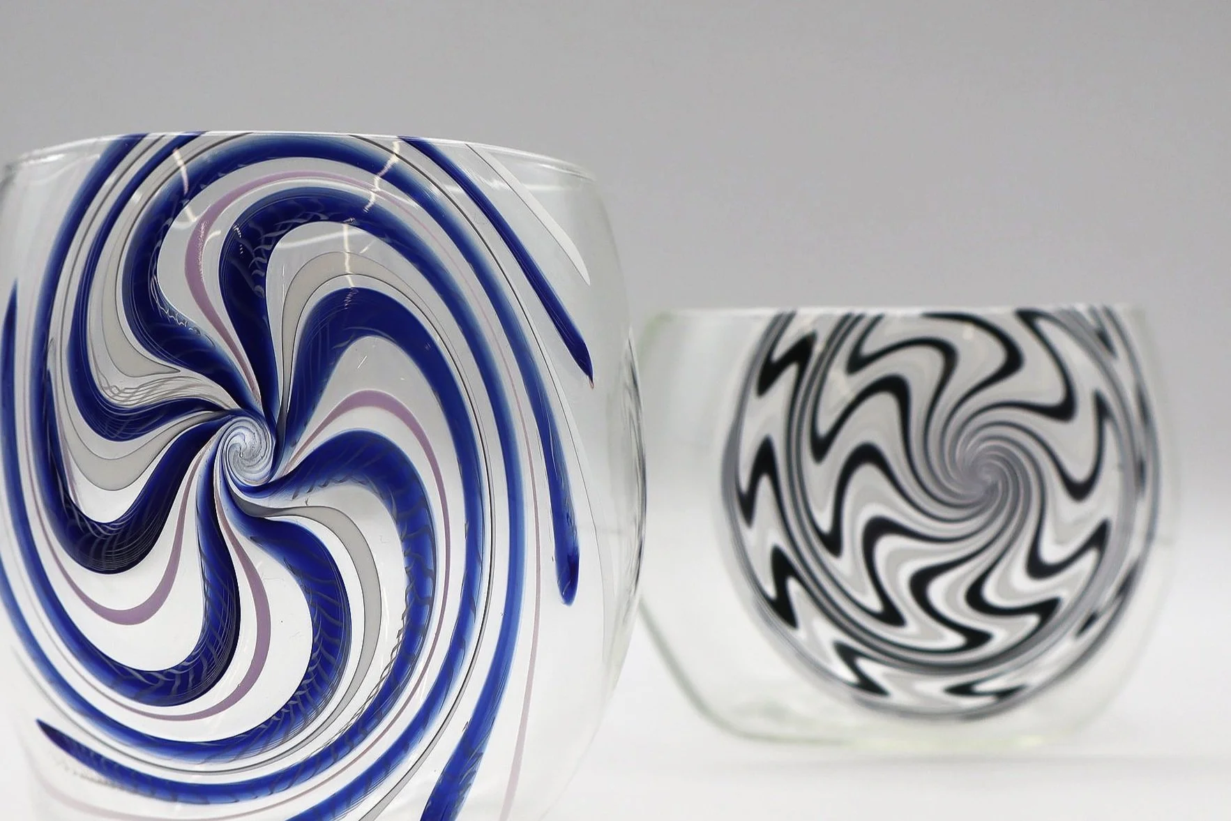 Due bicchieri di vetro artistici con motivi a spirale, uno con disegni blu e viola e l'altro con disegni neri e bianchi, posizionati uno accanto all'altro su uno sfondo neutro.