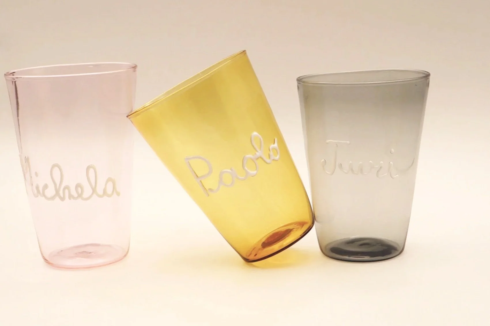 Tre bicchieri di vetro trasparente con nomi incisi: 'Michela', 'Rado', e 'Juri'. Uno di colore giallo inclinato.