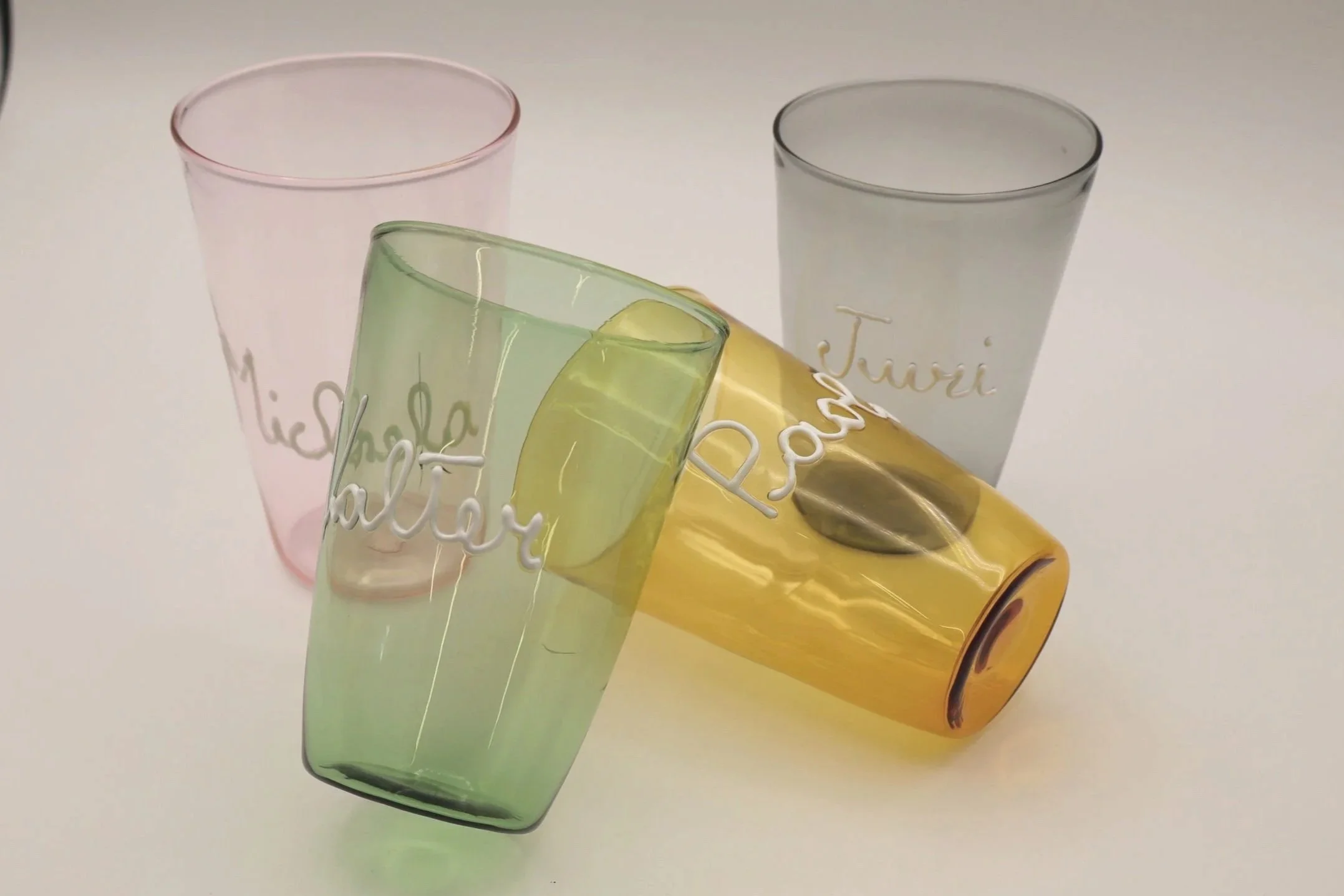 Quattro bicchieri di vetro colorati con scritte in italiano, uno di questi è giallo e si trova sopra gli altri, con una moneta al suo interno.