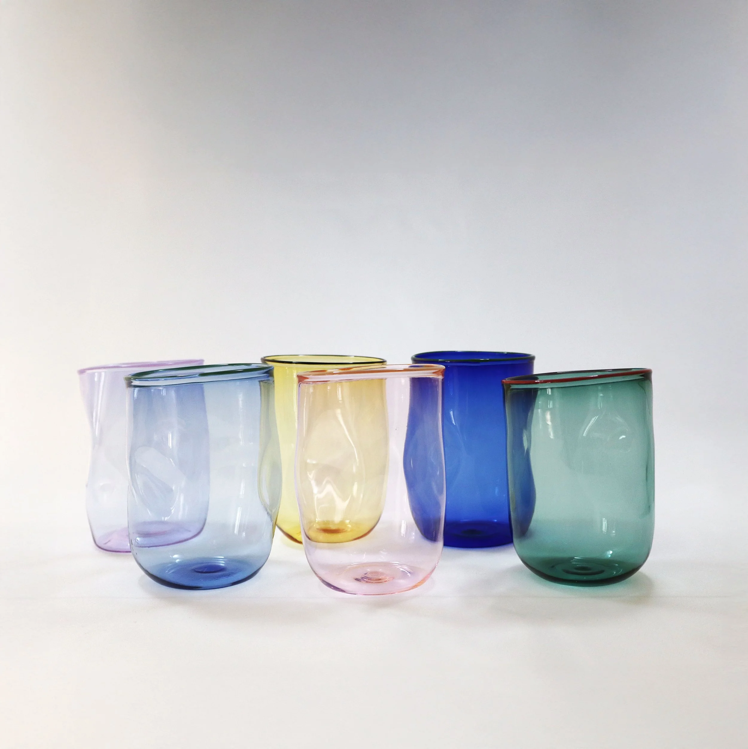 Set di sei bicchieri colorati in vetro trasparente, disposti su una superficie bianca di fronte a uno sfondo bianco.