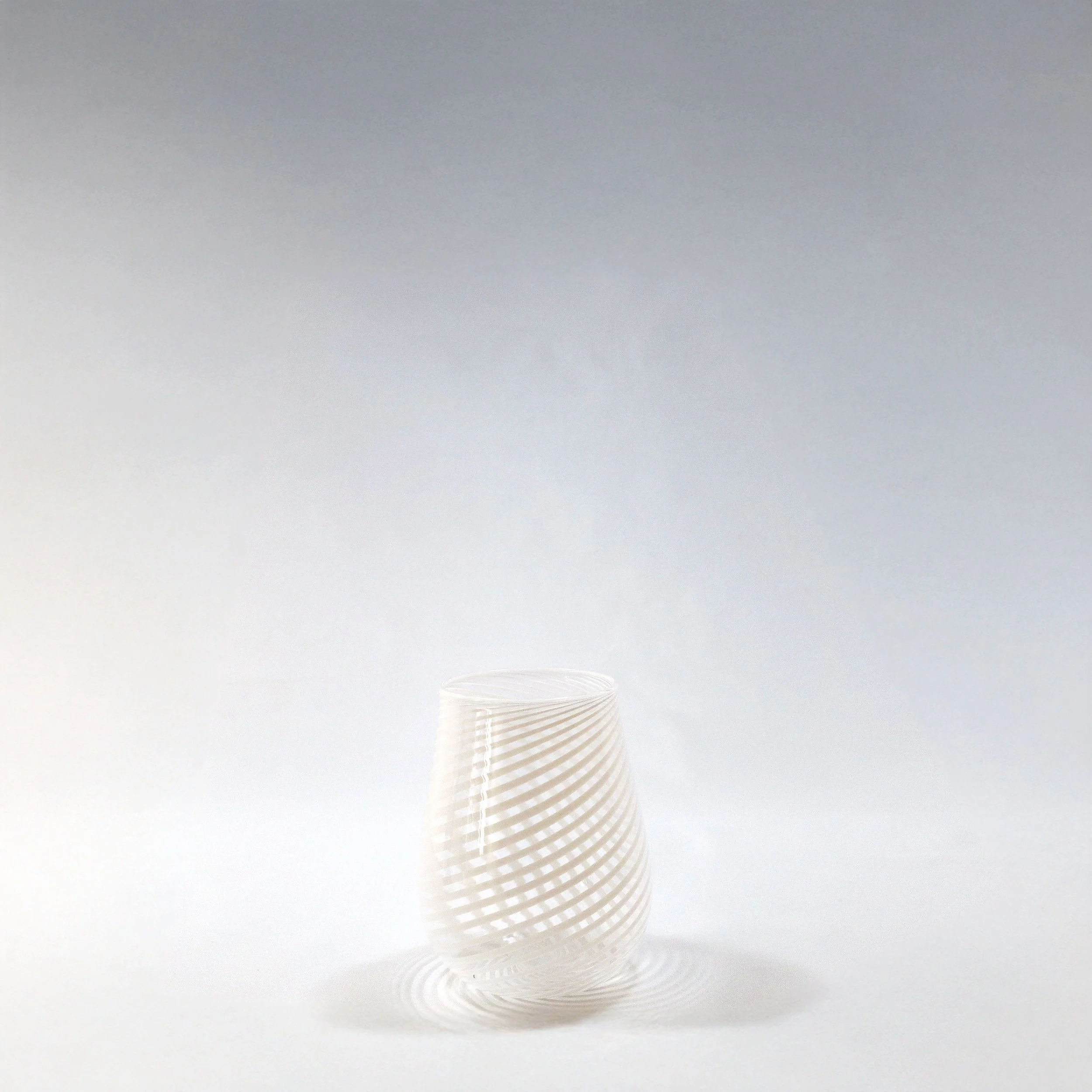 Bicchiere di vetro trasparente con un motivo a spirale bianco, posato su un piano bianco con uno sfondo bianco sfumato.