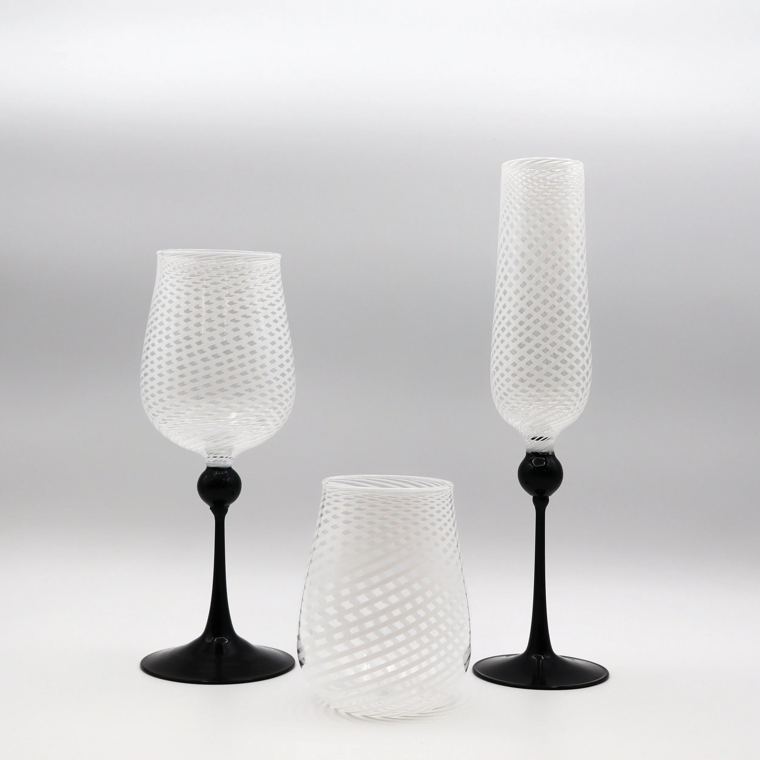 Tre bicchieri di vetro decorati con motivi a spirale, due con stelo nero e uno senza. Sono disposti su uno sfondo bianco.