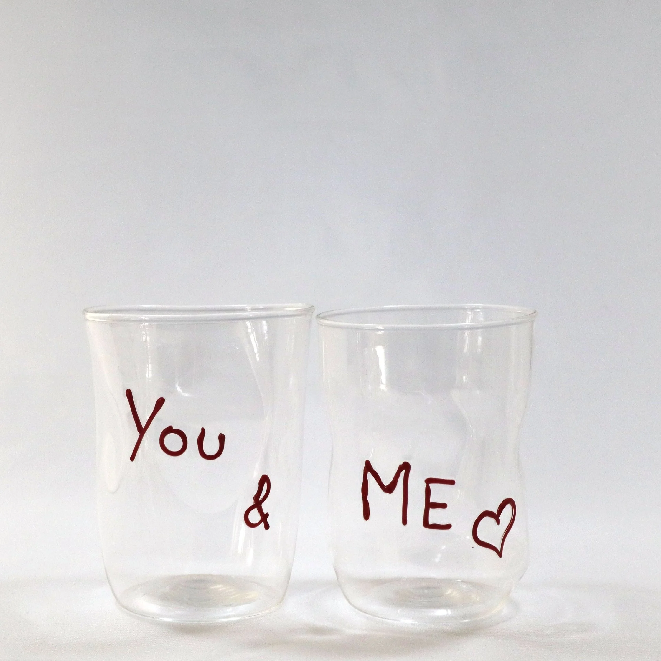Due bicchieri di plastica trasparenti con scritte rosse e cuori neri, uno con scritto 'You' e l'altro con scritto '& ME'