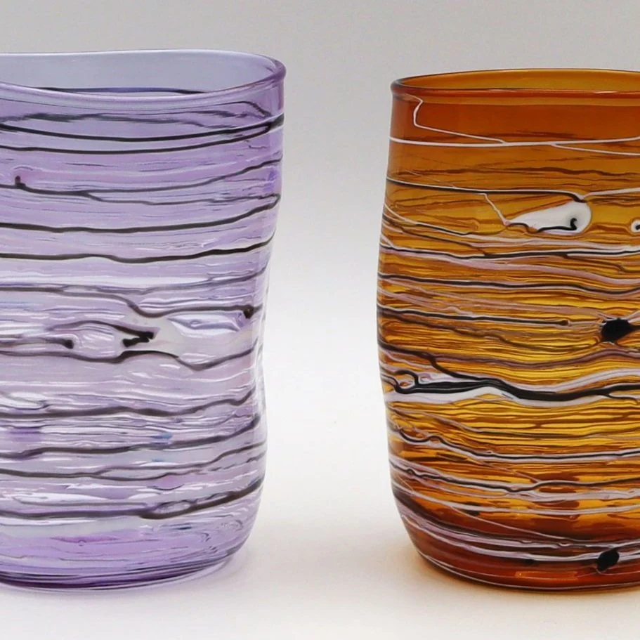 Due bicchieri di vetro colorati con motivi a righe fluide, uno viola e uno arancione, posizionati su uno sfondo bianco.