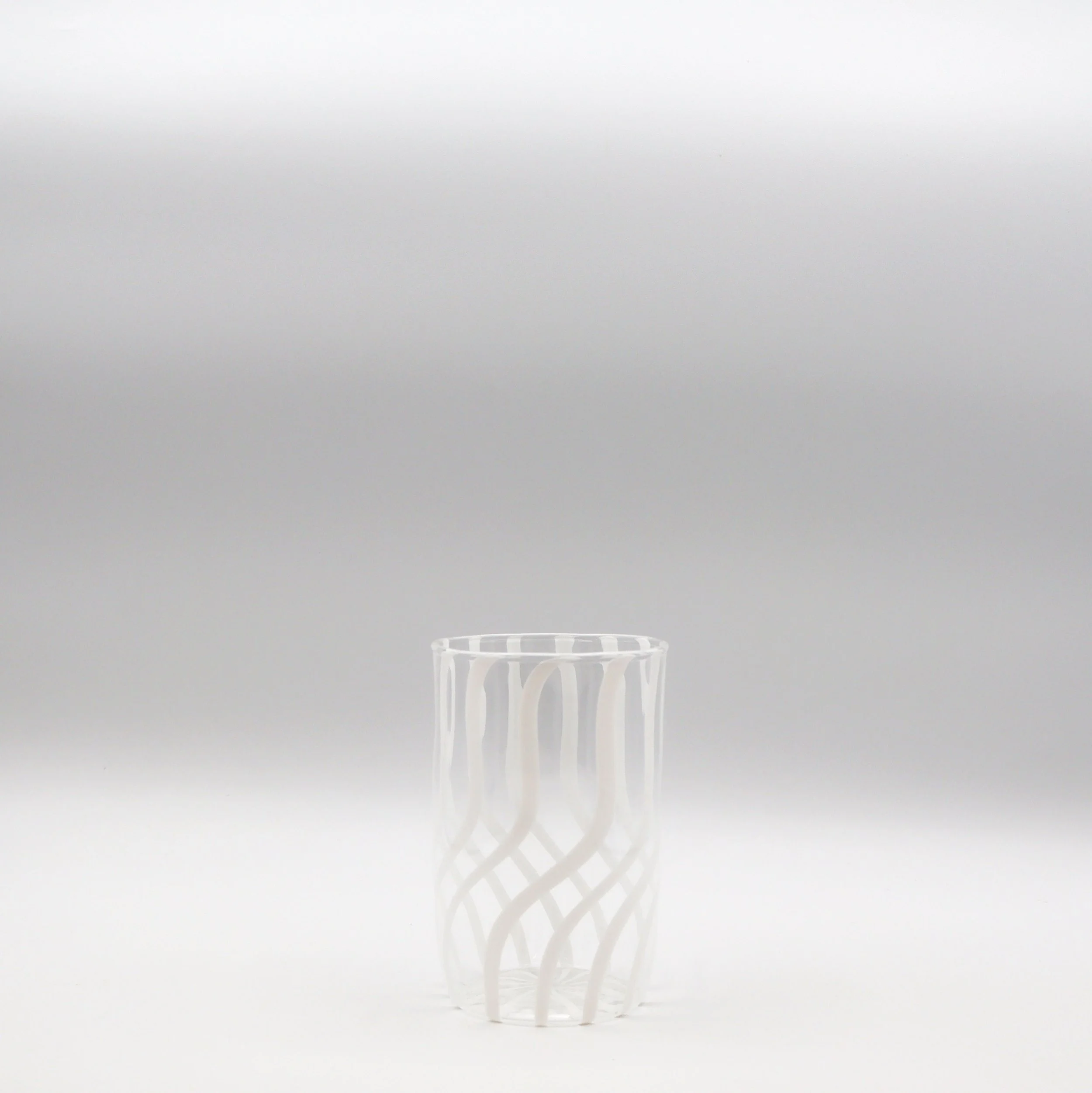 Vaso di vetro trasparente con motivo a spirale bianco, posizionato su uno sfondo grigio chiaro.