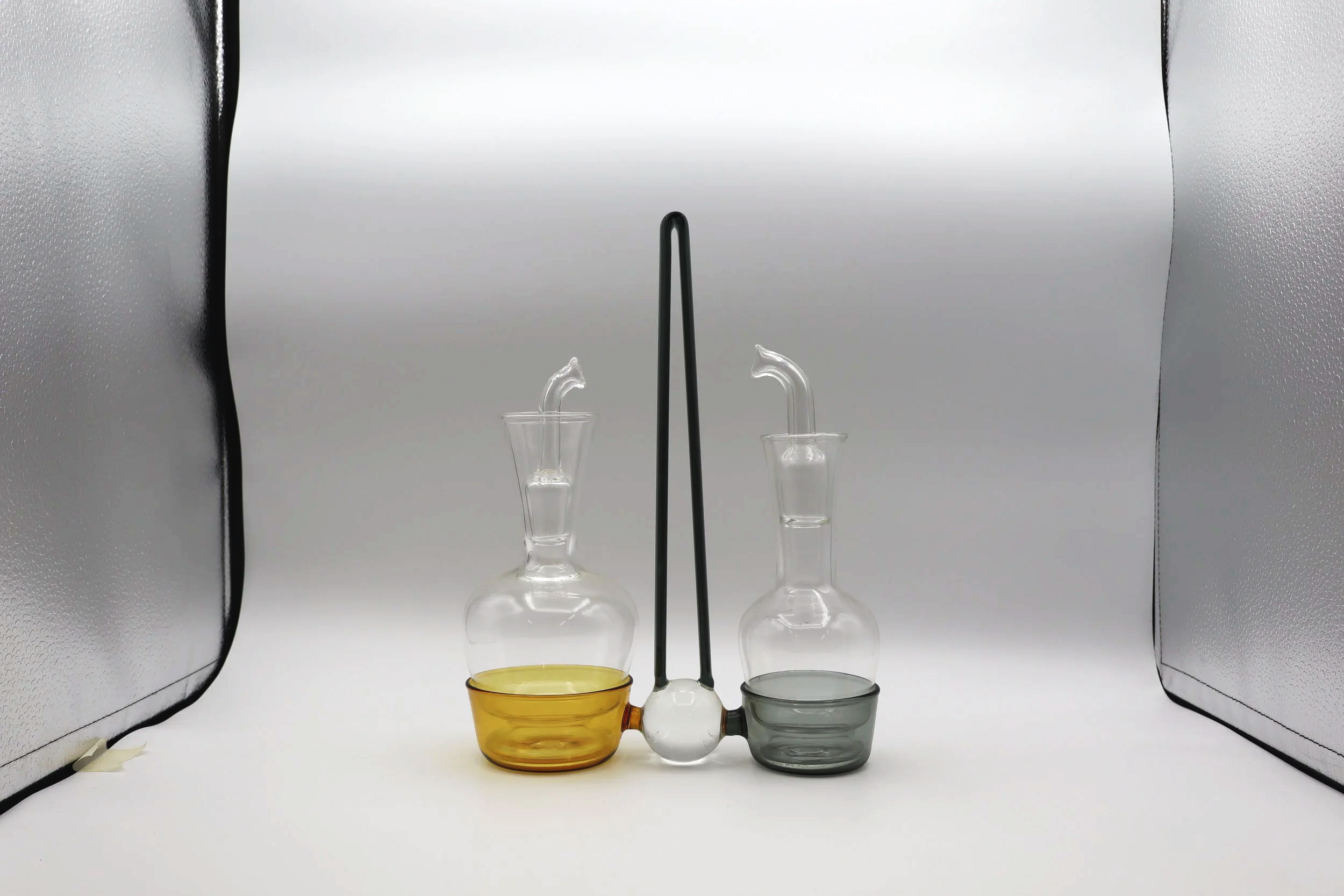 Due apparecchiature di laboratorio in vetro con tubi di raccordo, uno con un contenitore giallo e l'altro con uno grigio, collegati da un tubo e una sfera in vetro bianca al centro, in uno studio di fotografia con sfondo bianco