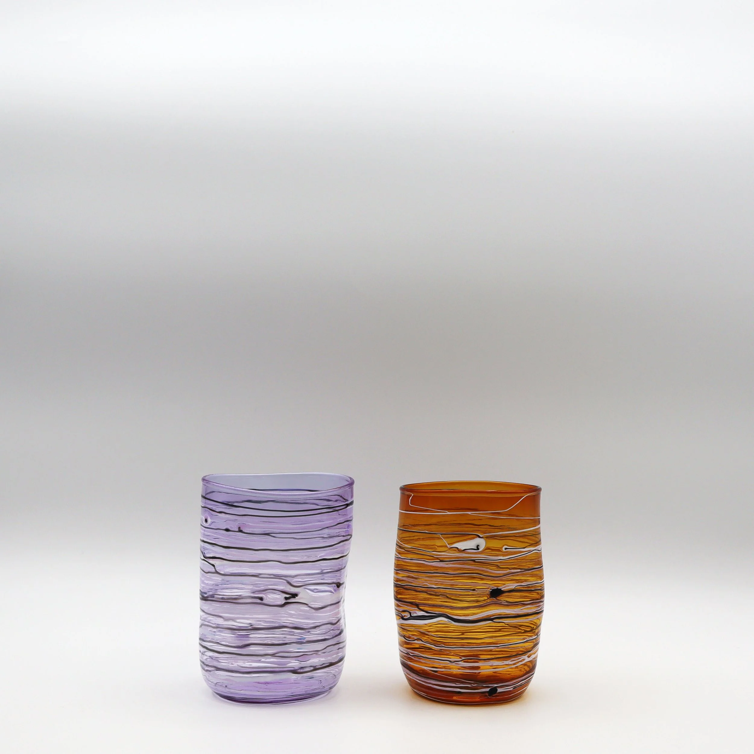 Due bicchieri di vetro, uno viola e uno arancione, con motivi di linee ondulate orizzontali, posti su uno sfondo bianco.