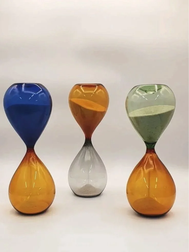 Tre clessidre di vetro colorate con sabbia di diversi colori, disposte in fila su uno sfondo bianco.