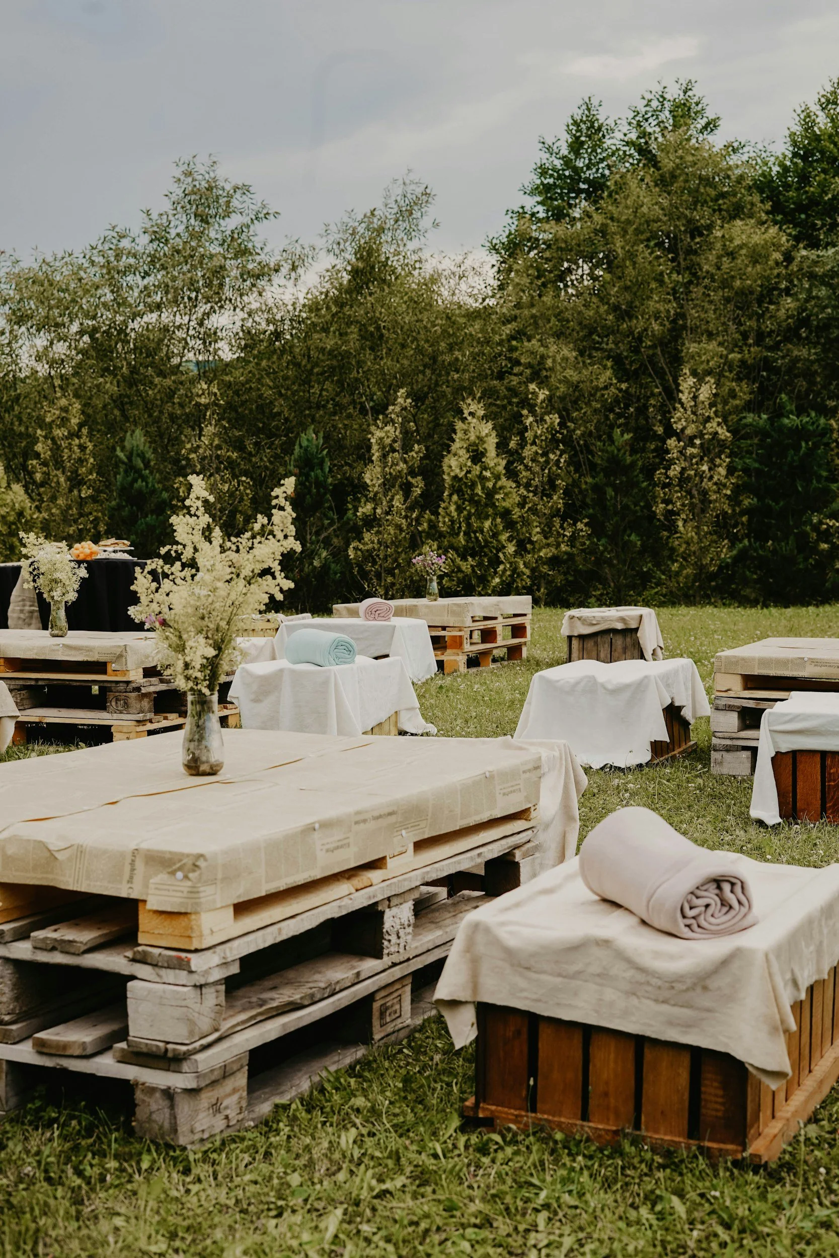 Outdoor rustic event setup with tables made from pallets, covered with white cloths, and decorated with vases of white flowers. Rolled towels are on some tables, and the background features trees and bushes under a cloudy sky.