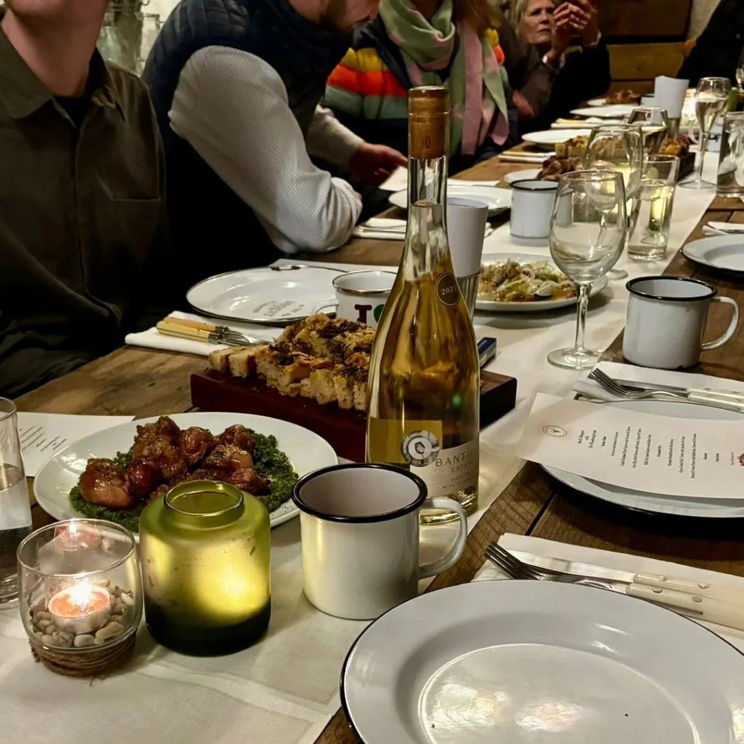 A table set on farm for a dining experience with various dishes, glasses, and a bottle of white wine, surrounded by people at the first Buck's Banquet.