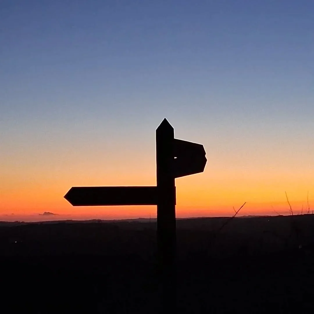 Silhouette of a directional signpost against a sunset sky with orange and blue hues, with distant hills on the horizon.