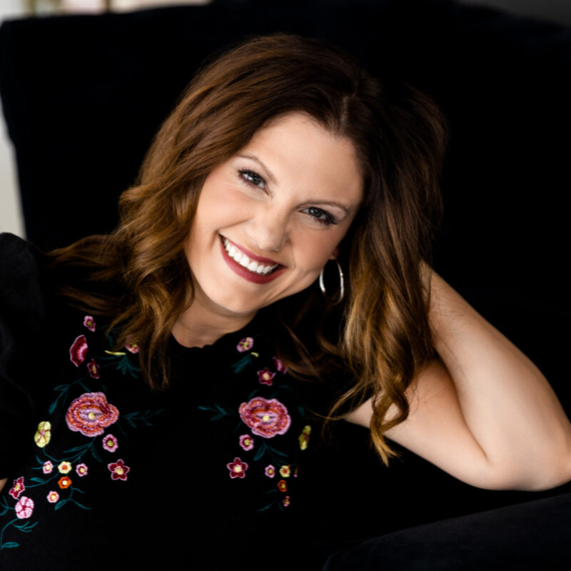 Smiling woman with shoulder-length brown hair, wearing a green short-sleeve top and dark jeans, sitting cross-legged against a white background.