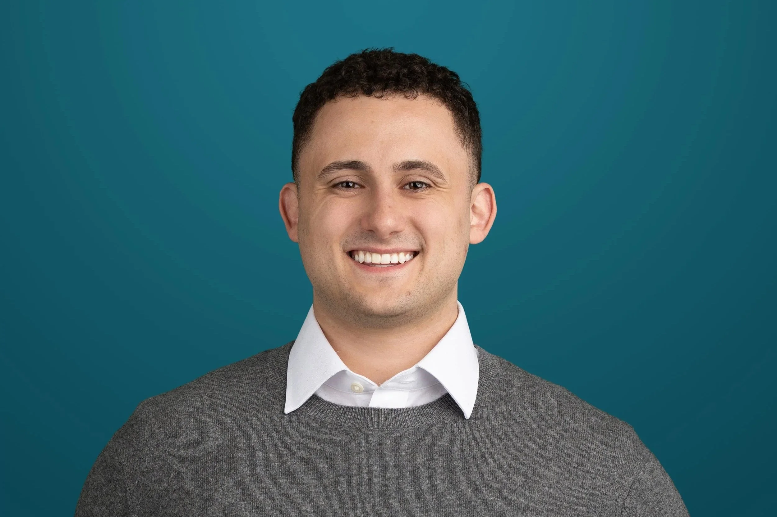 A young man with short dark hair, smiling, wearing a white collared shirt and a gray sweater, against a teal background.