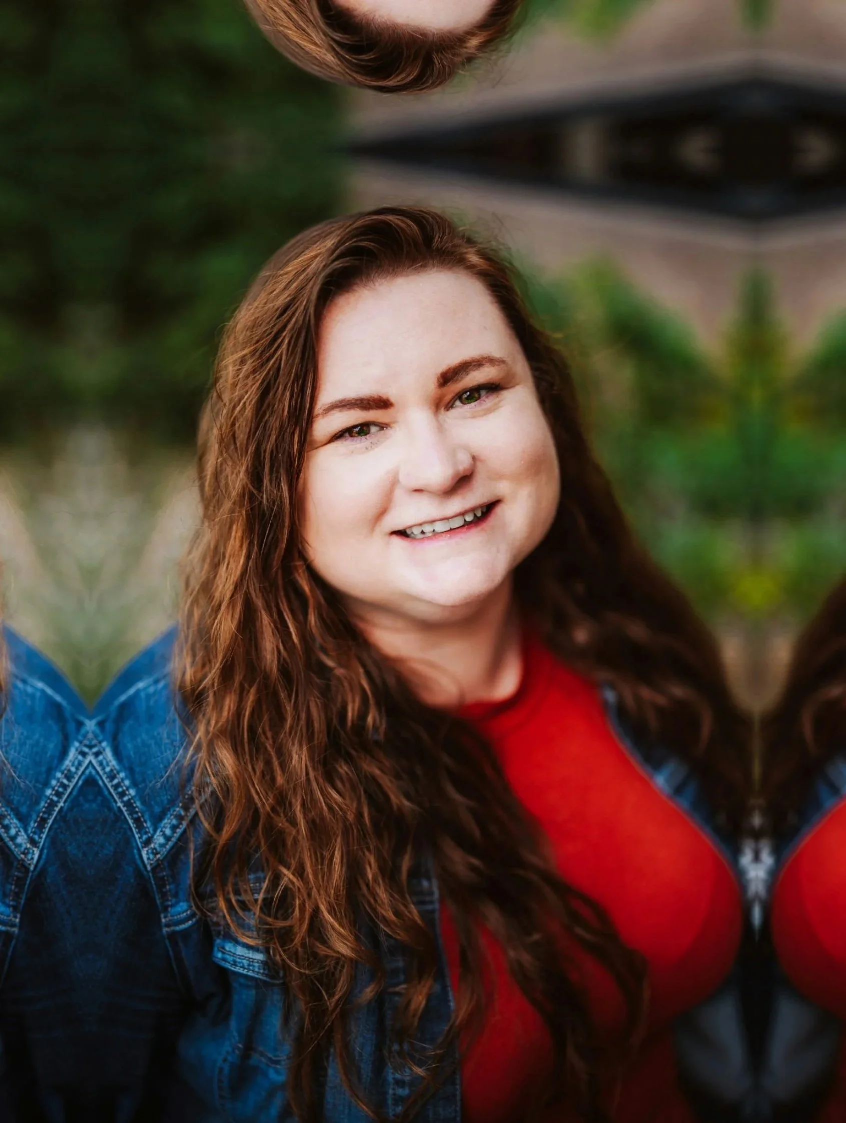 A close-up of a woman with long, wavy brown hair, smiling at the camera, outdoors with greenery in the background, wearing a red top and a denim jacket.