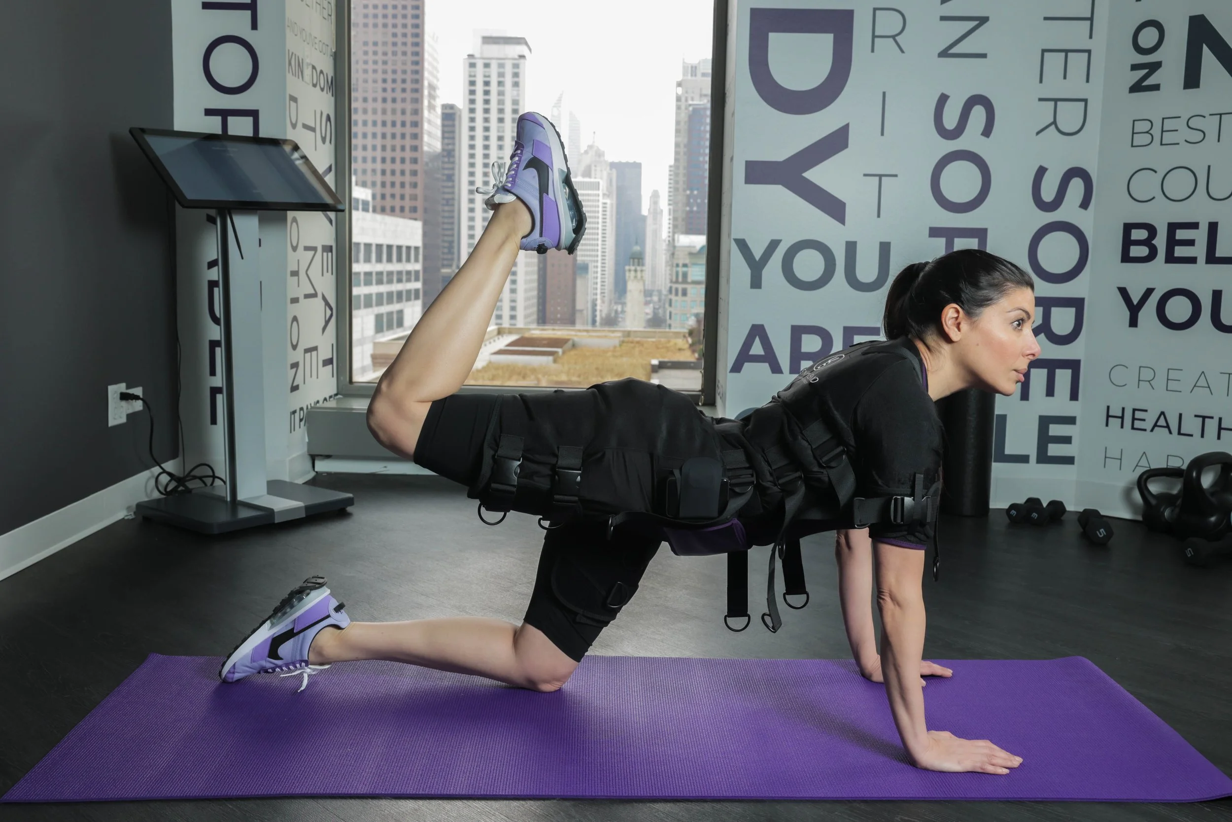 A woman in workout EMS (Electric Muscle Stimulation) suite performing a donkey kick exercise on a purple yoga mat in a gym with a Gold coast Chicago view outside the window.