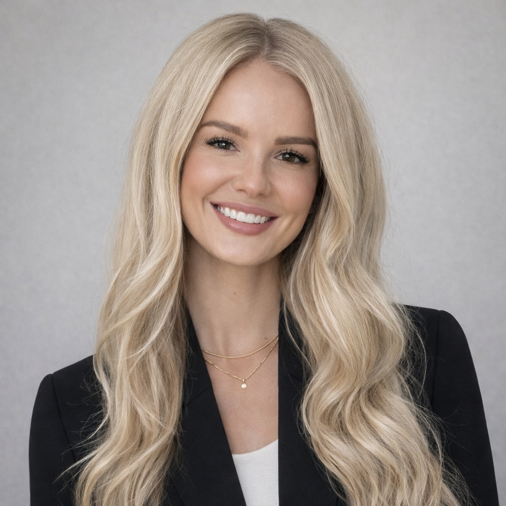 A young woman with long blonde hair smiling, wearing a black blazer and layered gold necklaces against a plain gray background.