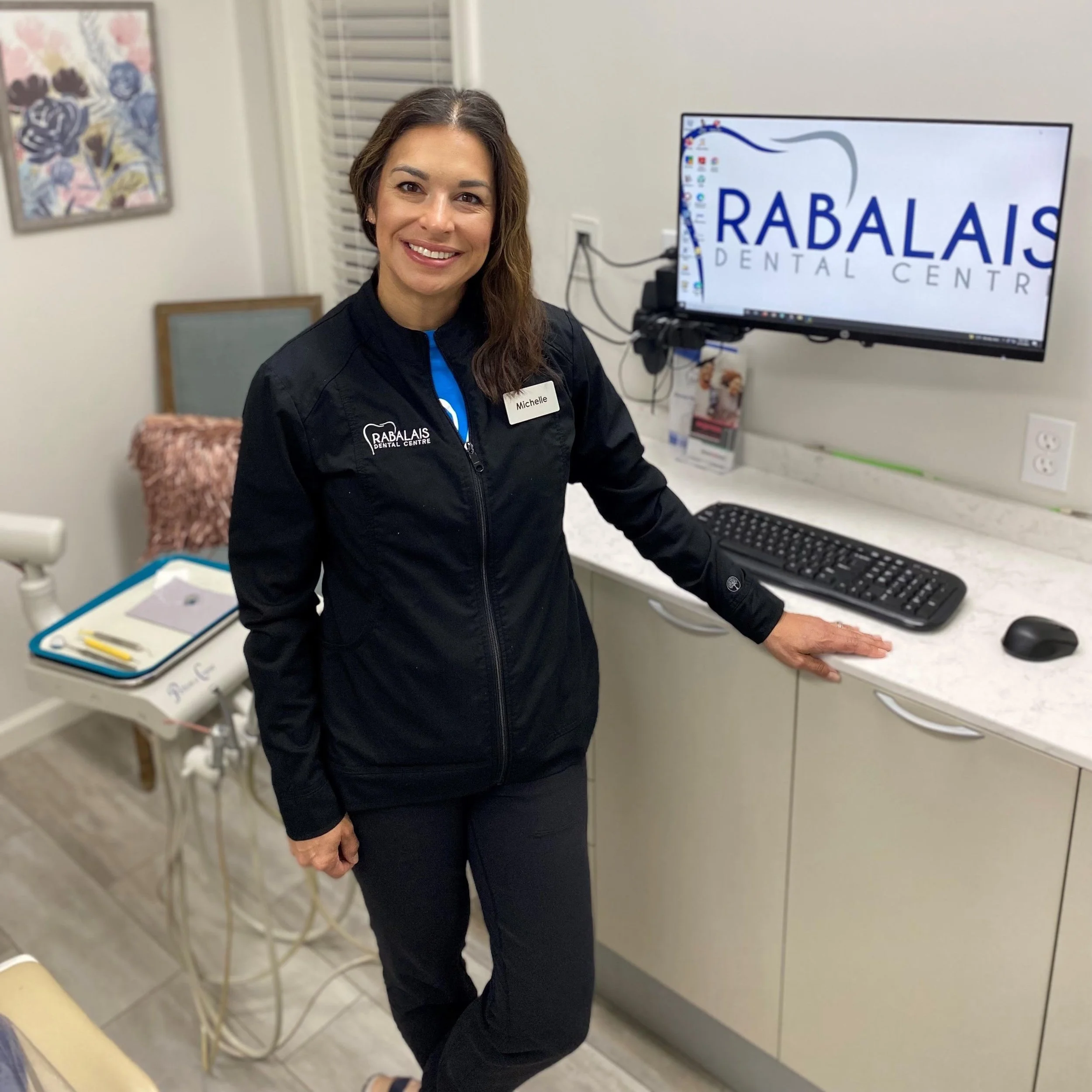 Dental office with a female staff member wearing a black jacket and name tag, standing near a computer displaying the Rabalais Dental Center logo.