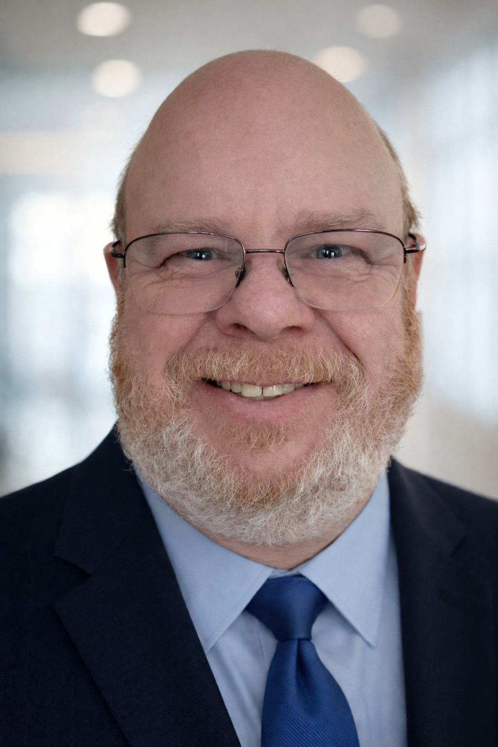 Close-up of a smiling man with glasses, a beard, and a bald head, wearing a suit and tie.