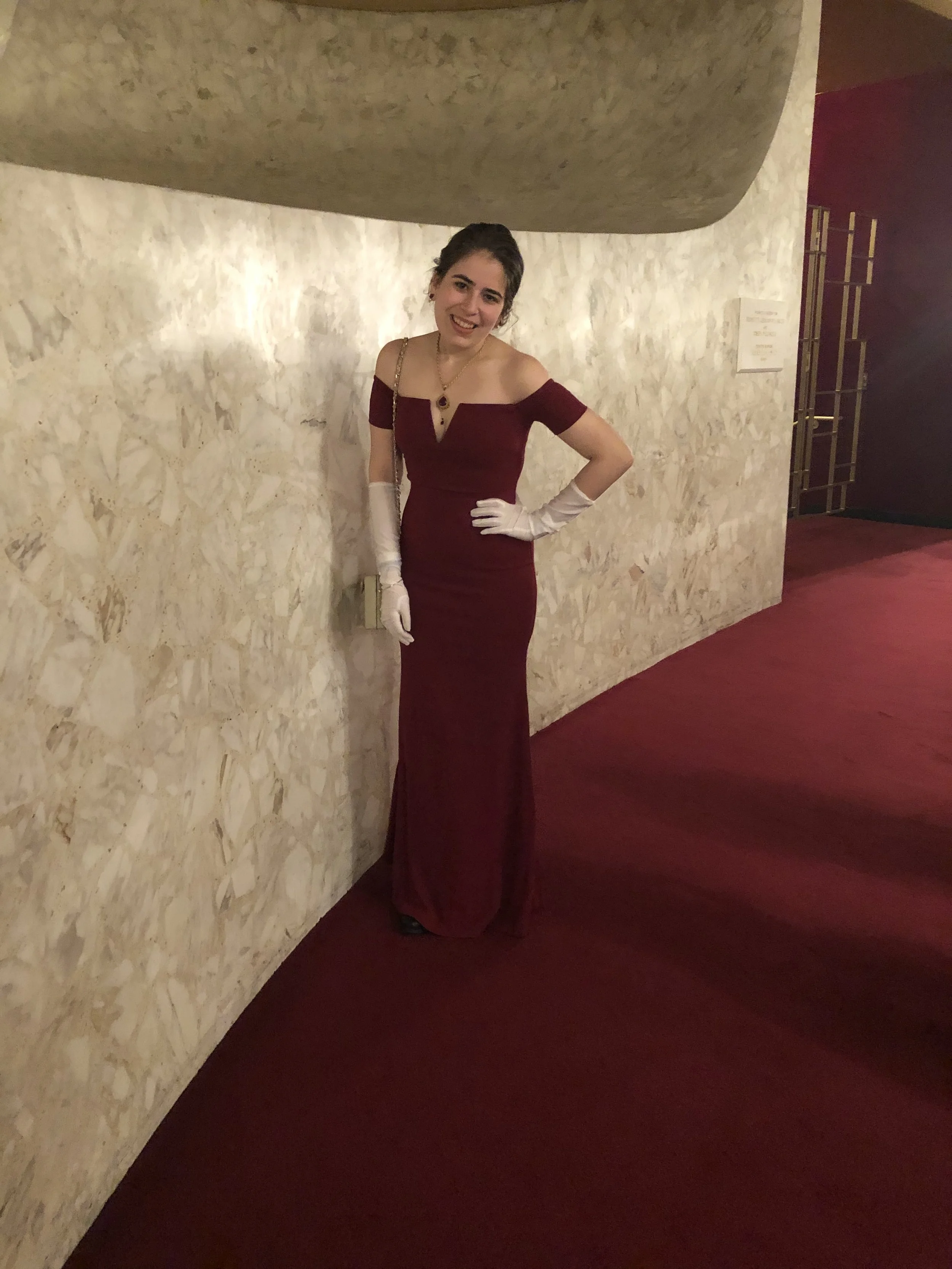 Person wearing a burgundy evening gown and white gloves, standing in a hallway with marble walls and red carpet.