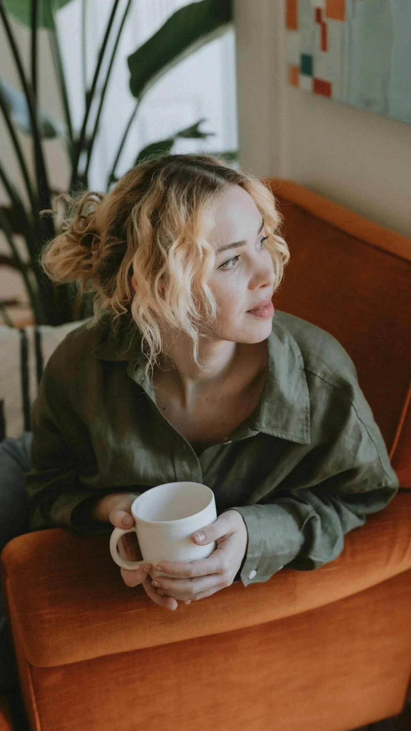 Woman with curly blonde hair holding a white coffee mug at a wooden café table