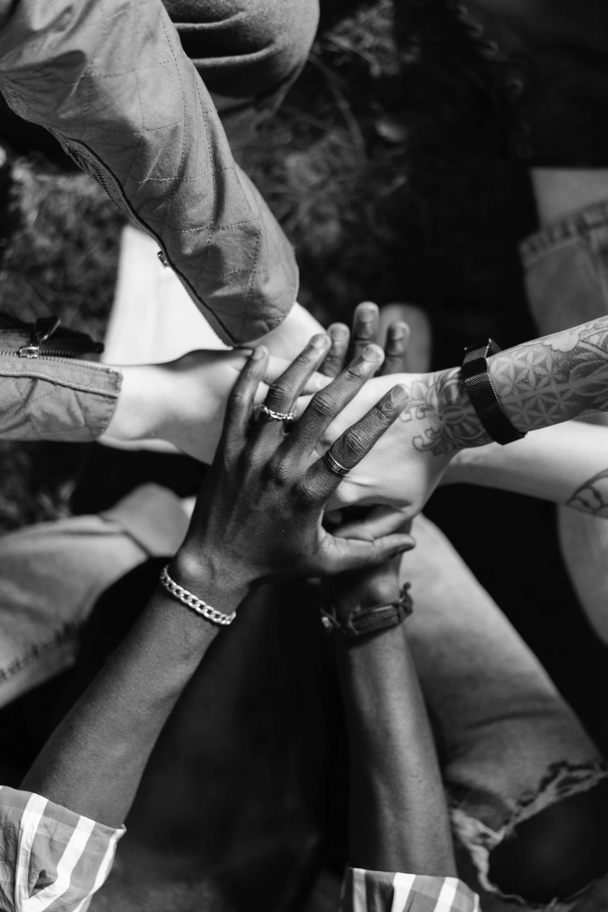Multiple hands from diverse individuals stacked together showing unity and support, with tattoos, jewelry, and varied clothing in black and white.