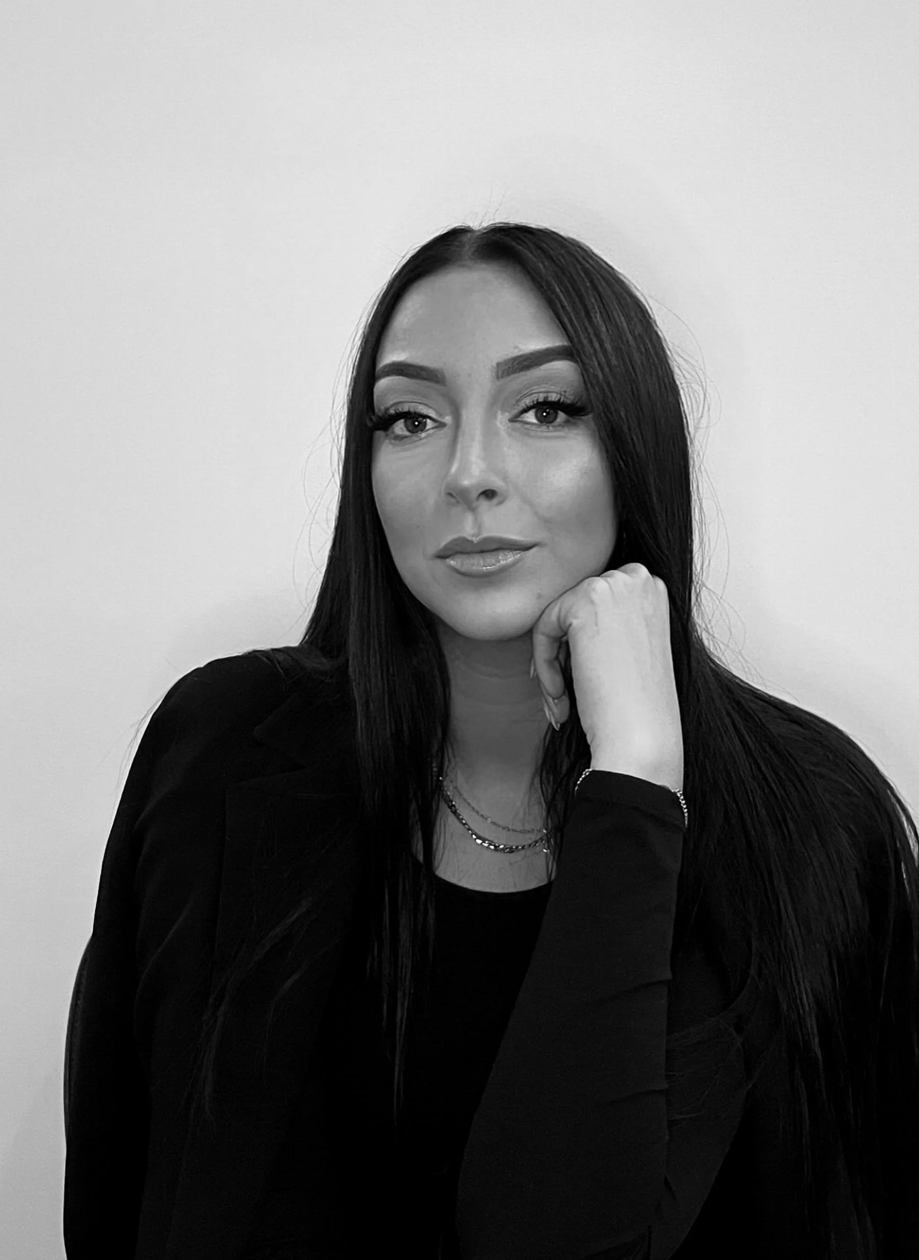 Black and white portrait of a woman with long dark hair, wearing a black top, resting her chin on her hand, against a plain background.
