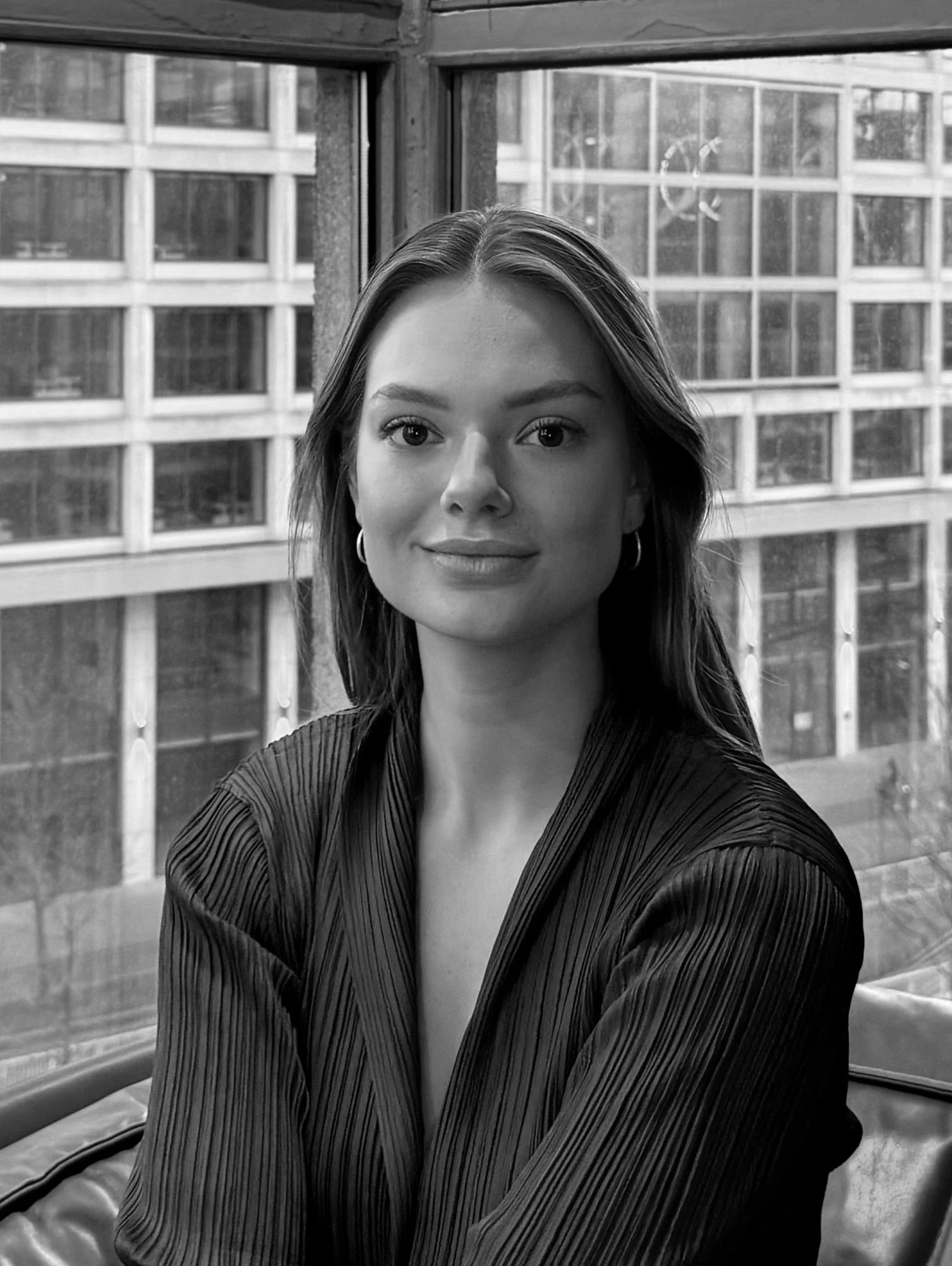 A young woman with long hair, wearing earrings and a pleated blouse, sitting in front of a large window with a cityscape of modern buildings in the background.