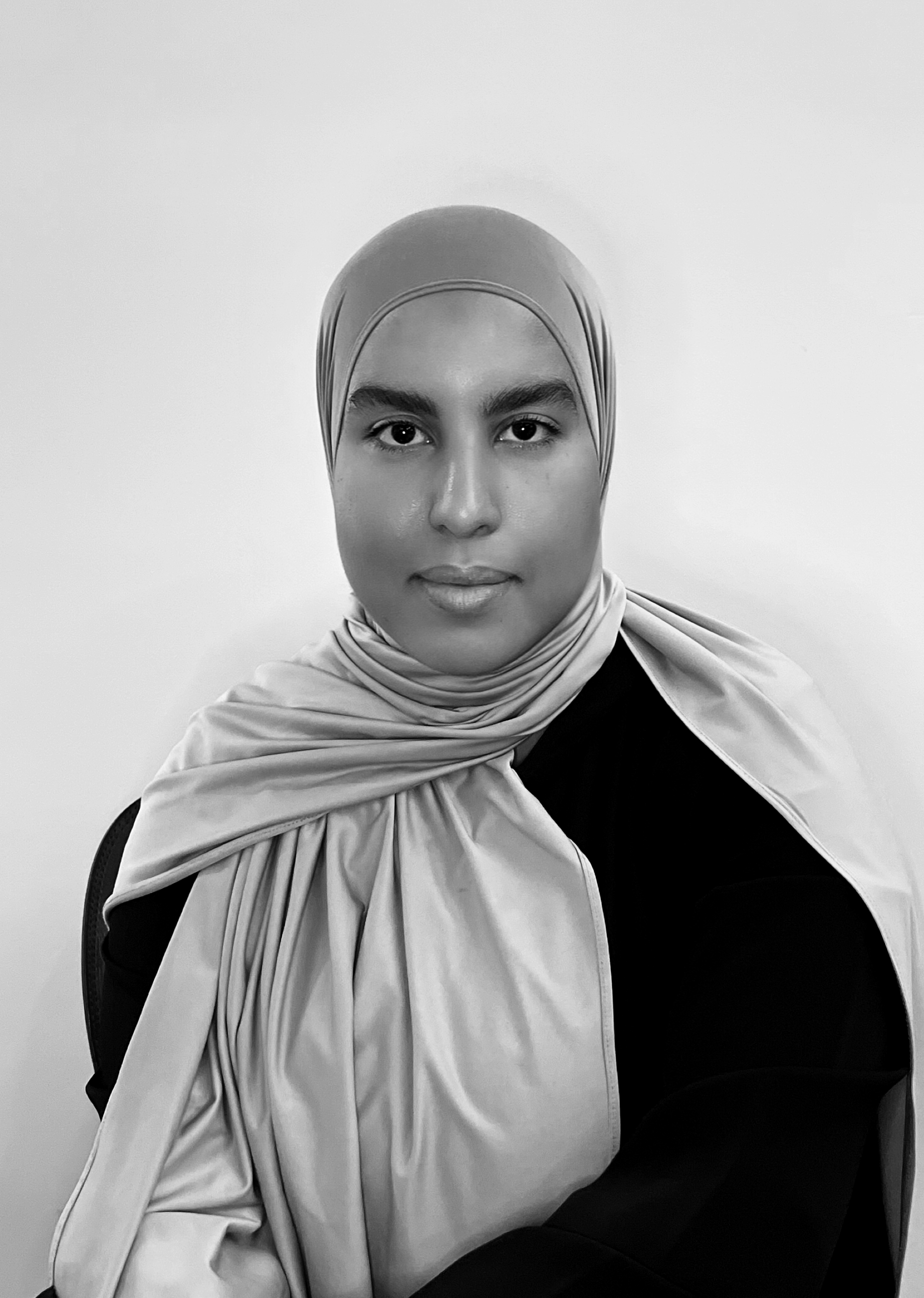 A woman wearing a hijab and a dark top, sitting against a plain light background, looking directly at the camera.
